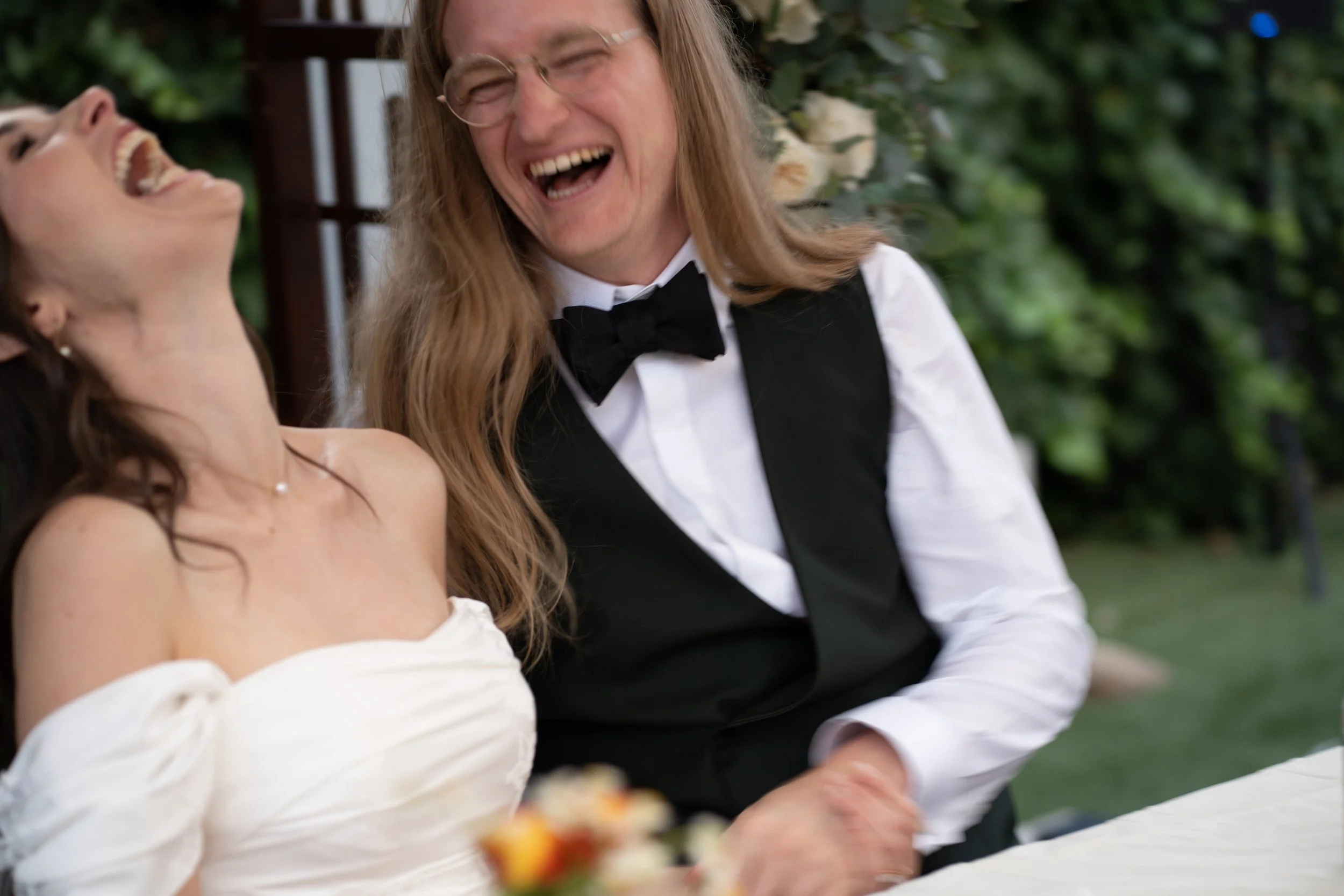 Two women laughing and enjoying a moment together outdoors, one wearing a white off-shoulder dress and the other in a black tuxedo with a bow tie, surrounded by greenery.
