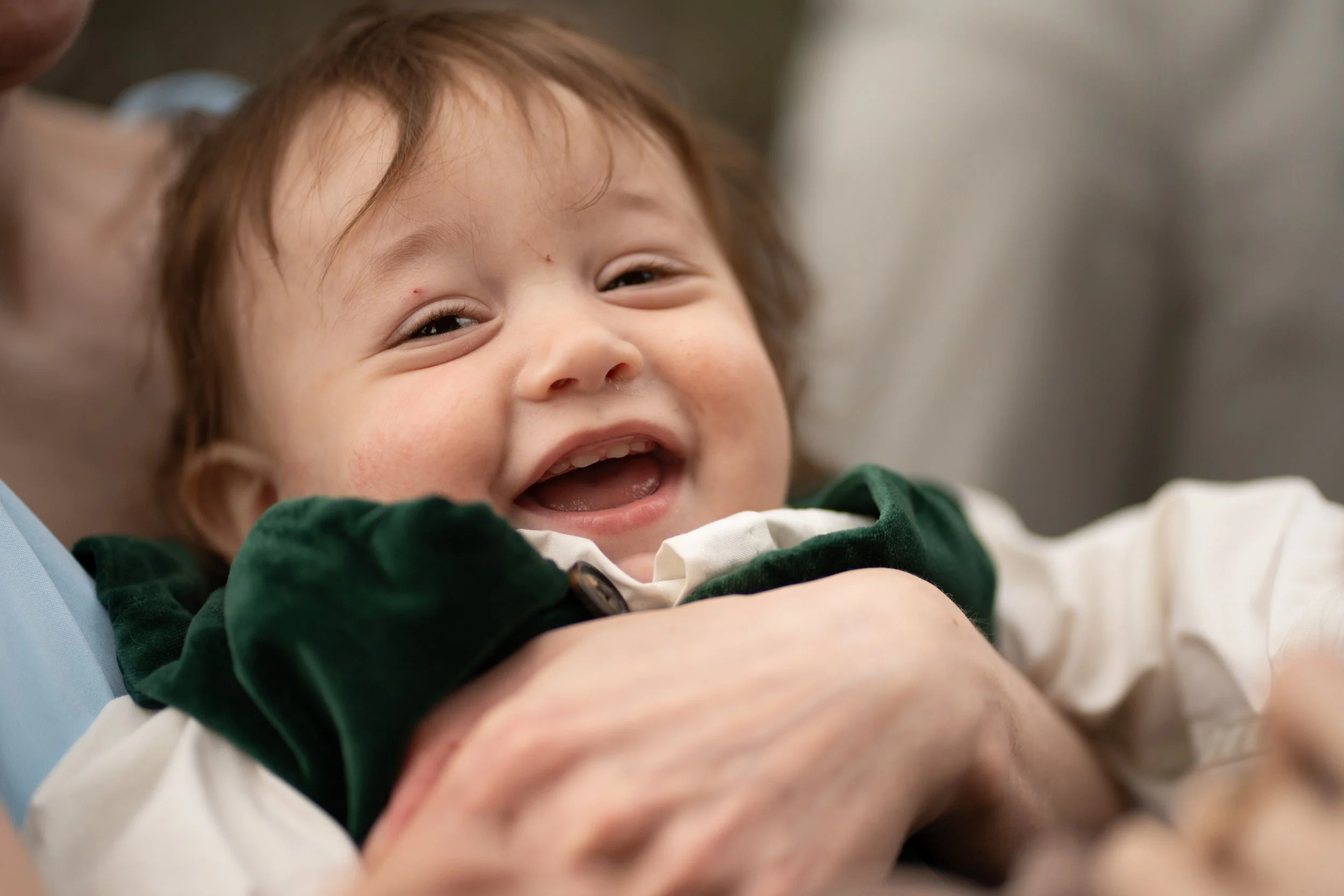 A smiling young child with brown hair lying in someone's arms, wearing a green and white shirt, with visible small red marks or pimples on their face.