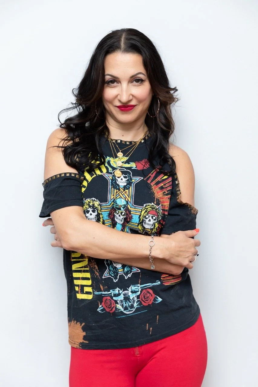 A woman with long dark hair, wearing a black Guns N' Roses t-shirt and red pants, standing against a plain white wall.