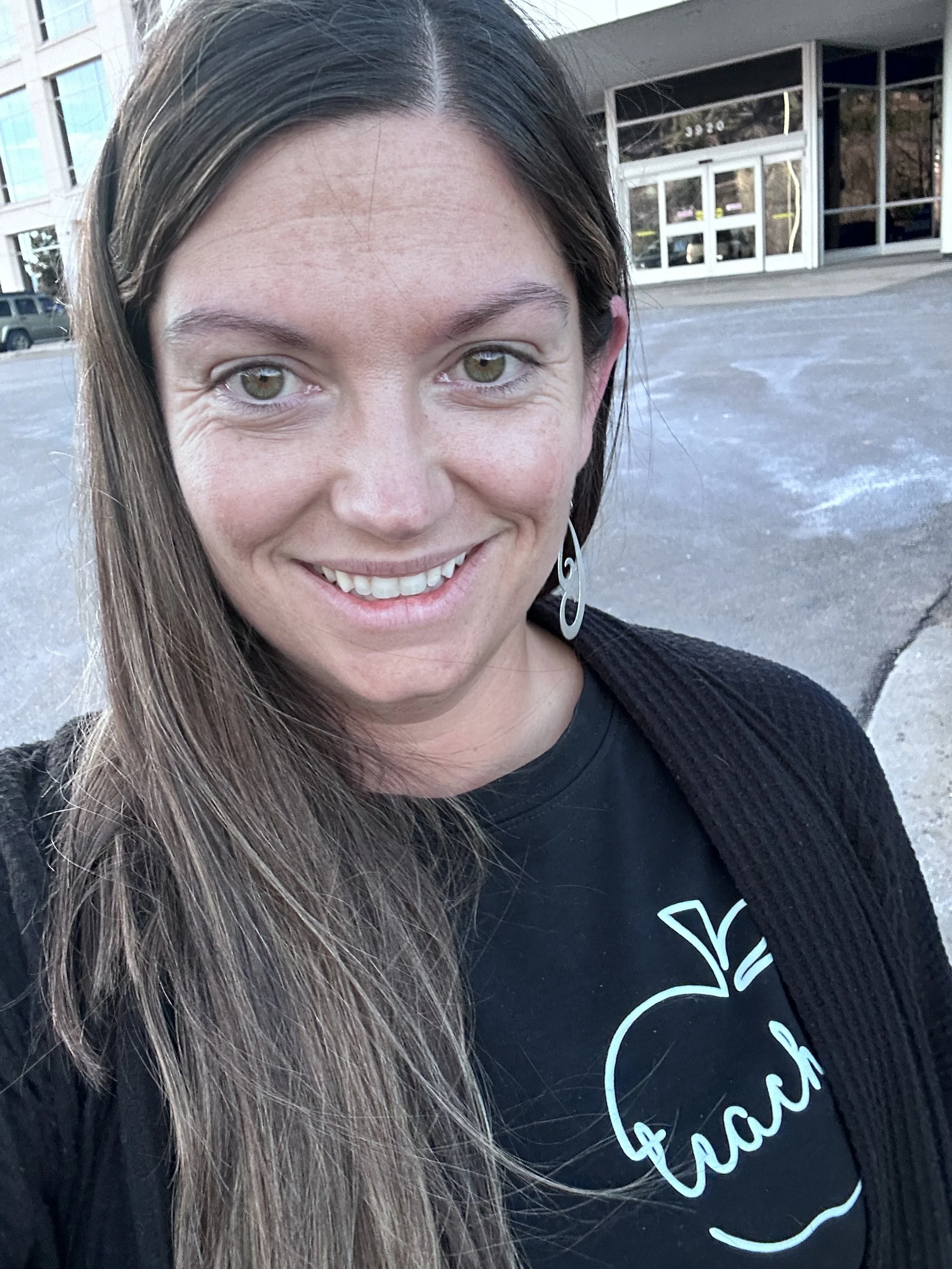 A smiling woman with long brown hair taking a selfie outdoors, wearing a black top with a white apple logo and the word 'eat' inside, and a black cardigan, with buildings and a parking lot in the background.