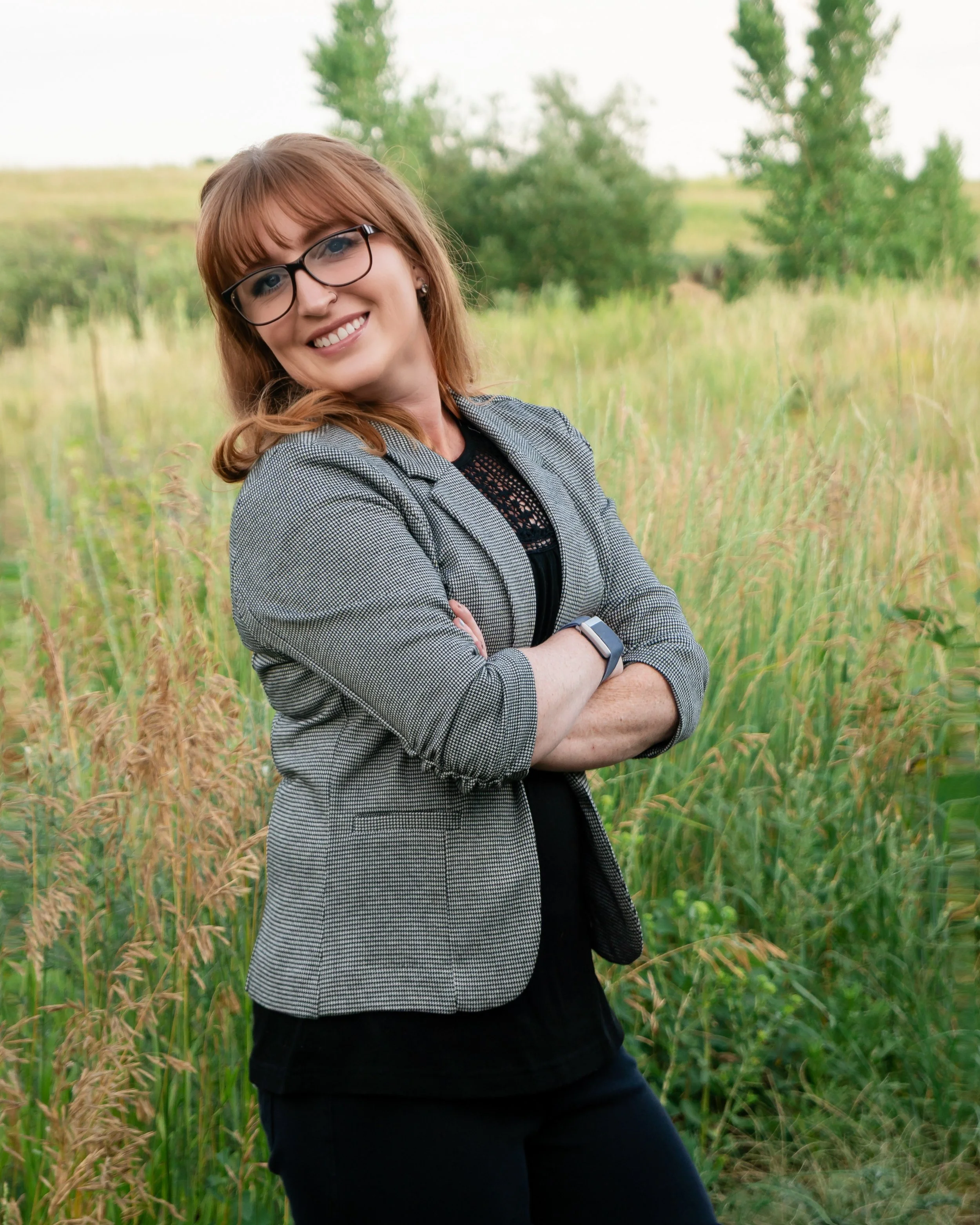 A woman with glasses and red hair smiling, standing with arms crossed in a field of tall grass and trees.