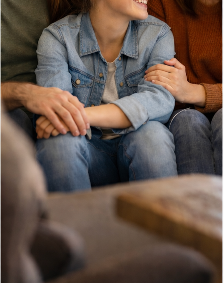 People sitting closely together with a woman in a denim jacket smiling, holding hands, and hugging a person wearing a rust-colored sweater.