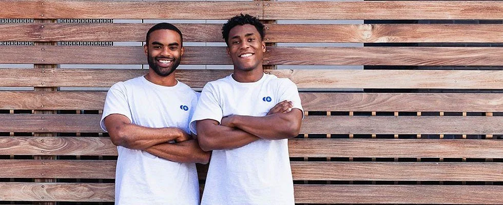 Two young black men smiling against a wooden slat wall.