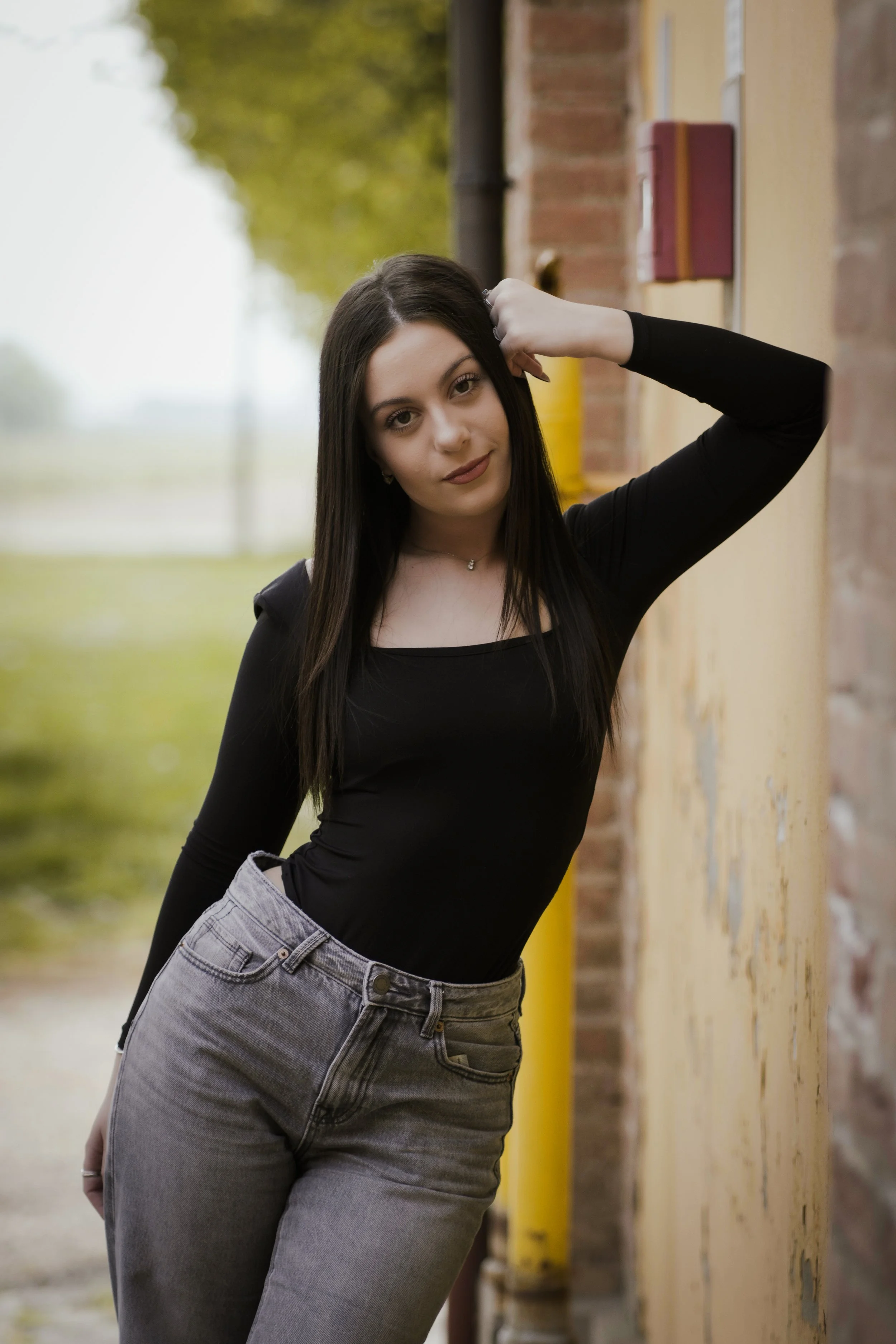 Ragazza con capelli corti neri, indossa una maglietta nera e pantaloni a vita alta grigi, posa vicino a un muro con sfondo naturale e alberi.