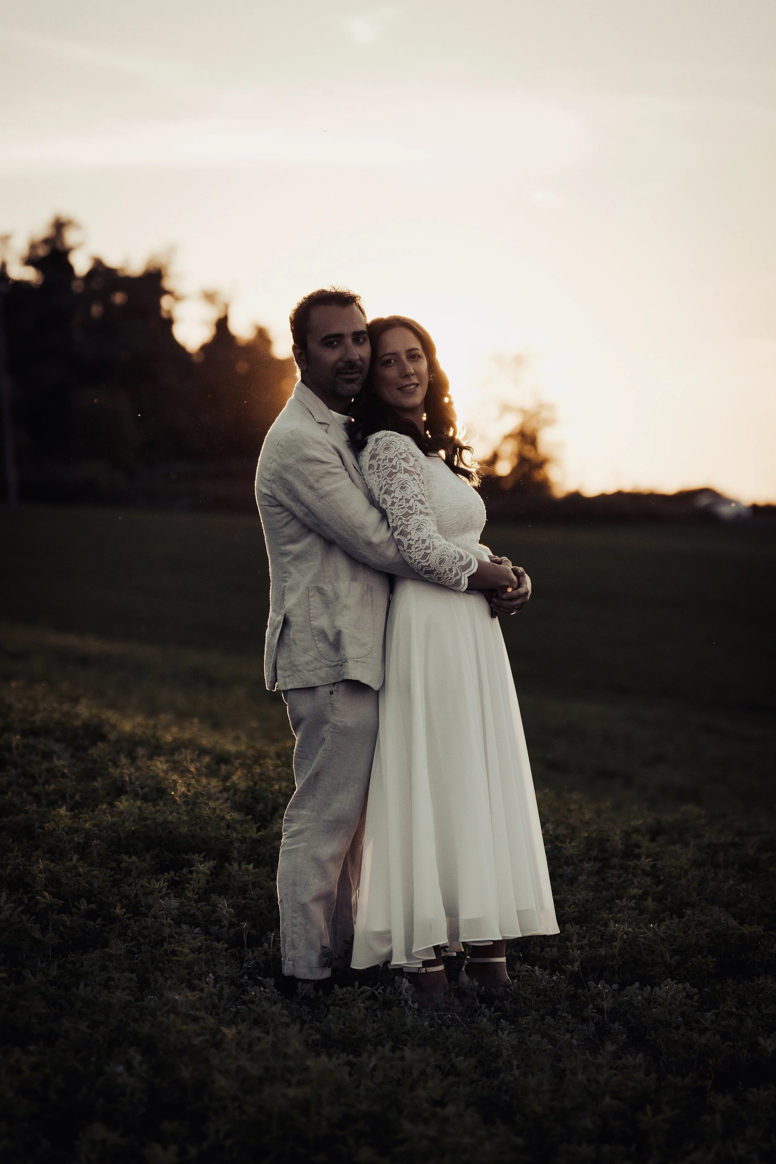 Una coppia felice e innamorata si abbraccia durante il tramonto in un prato verde, lei indossa un abito bianco con maniche di pizzo, lui un completo chiaro.