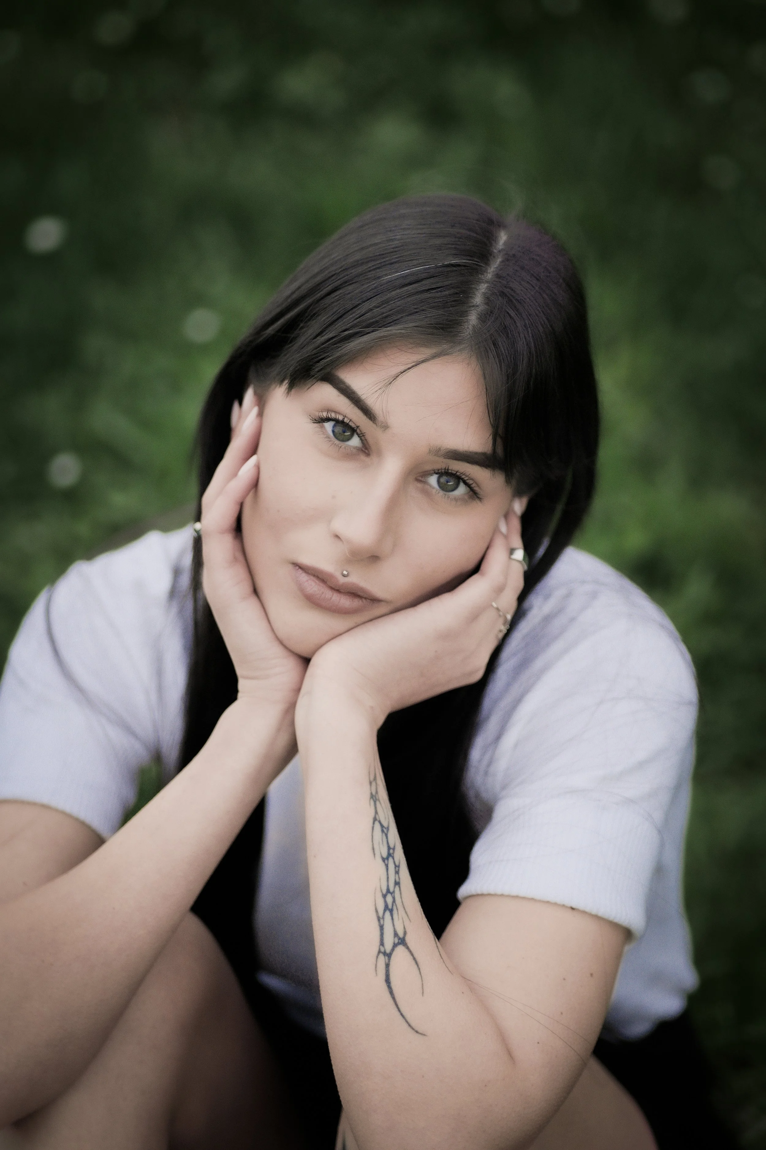 Ragazza con capelli neri, occhi azzurri, seduta sull'erba verde, con tatuaggio sul braccio e piercing alle labbra, che guarda verso la fotocamera.