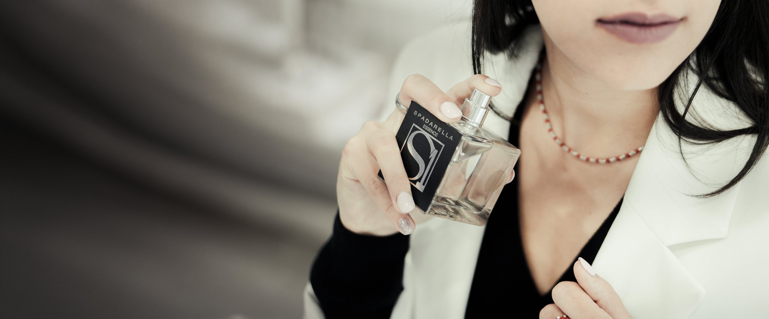 Donna con abito bianco e collana di perline rossa e bianca tiene un flacone di profumo trasparente con etichetta nera e bianca.