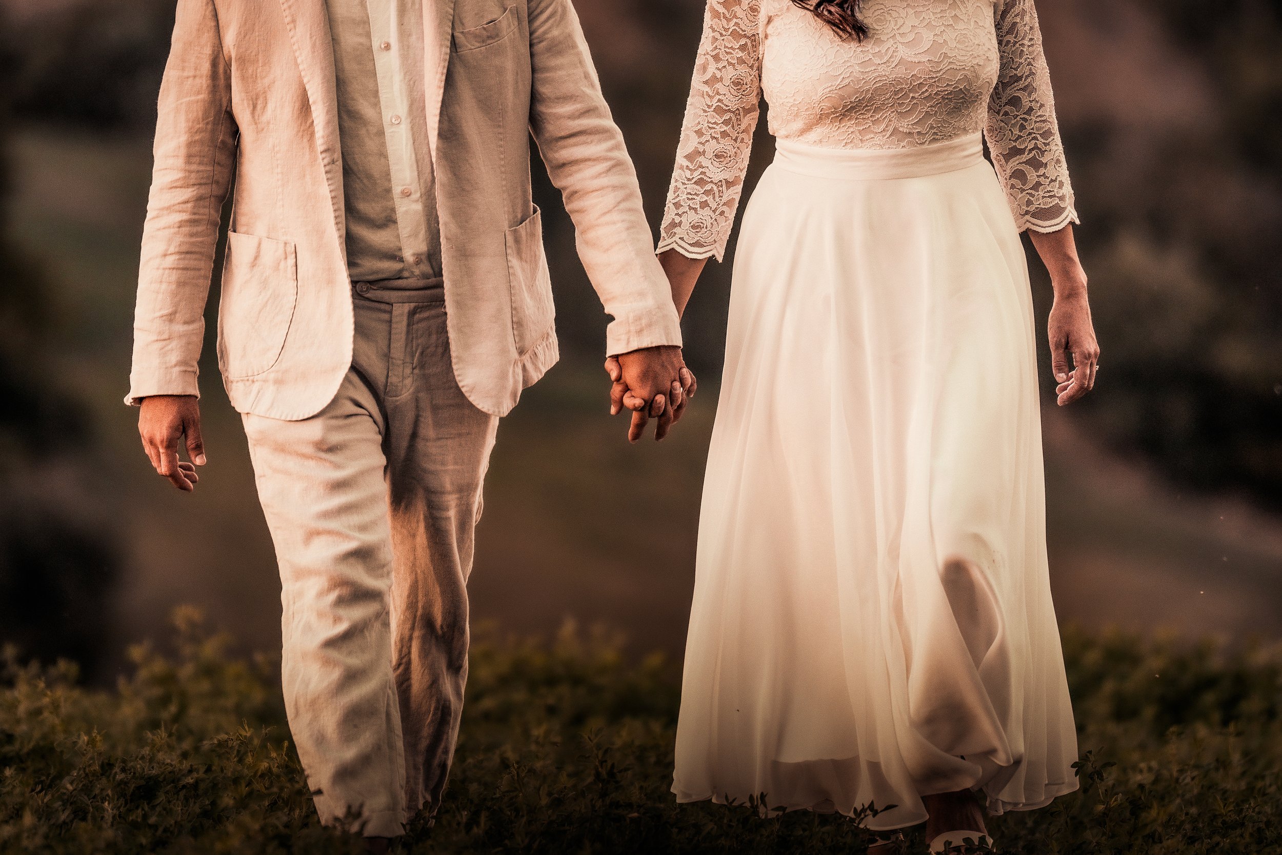 Un uomo e una donna che camminano mano nella mano all'aperto, lui in abito chiaro e lei in abito da sposa in pizzo, ambientazione al tramonto.