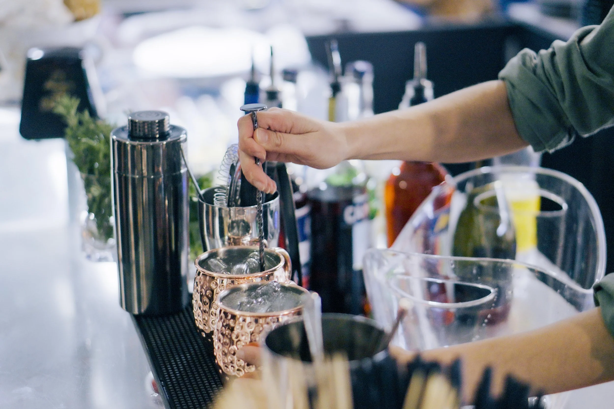Una mano sta preparando un cocktail con una cannuccia in un bar, con vari strumenti e bicchieri sul bancone.