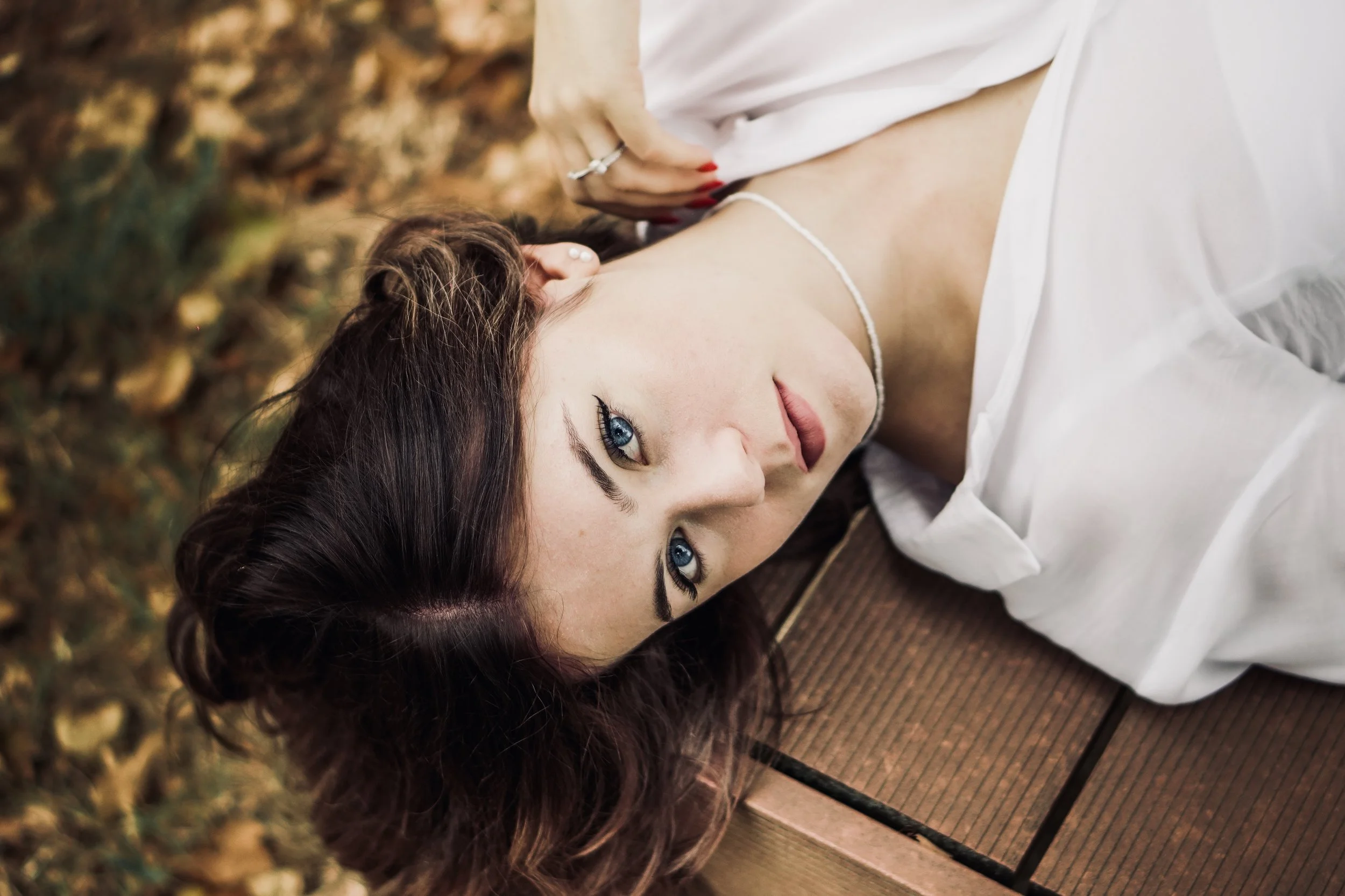 Donna con occhi azzurri e capelli ricci, sdraiata su un'area di legno, indossa una camicia bianca e un semplice collier.