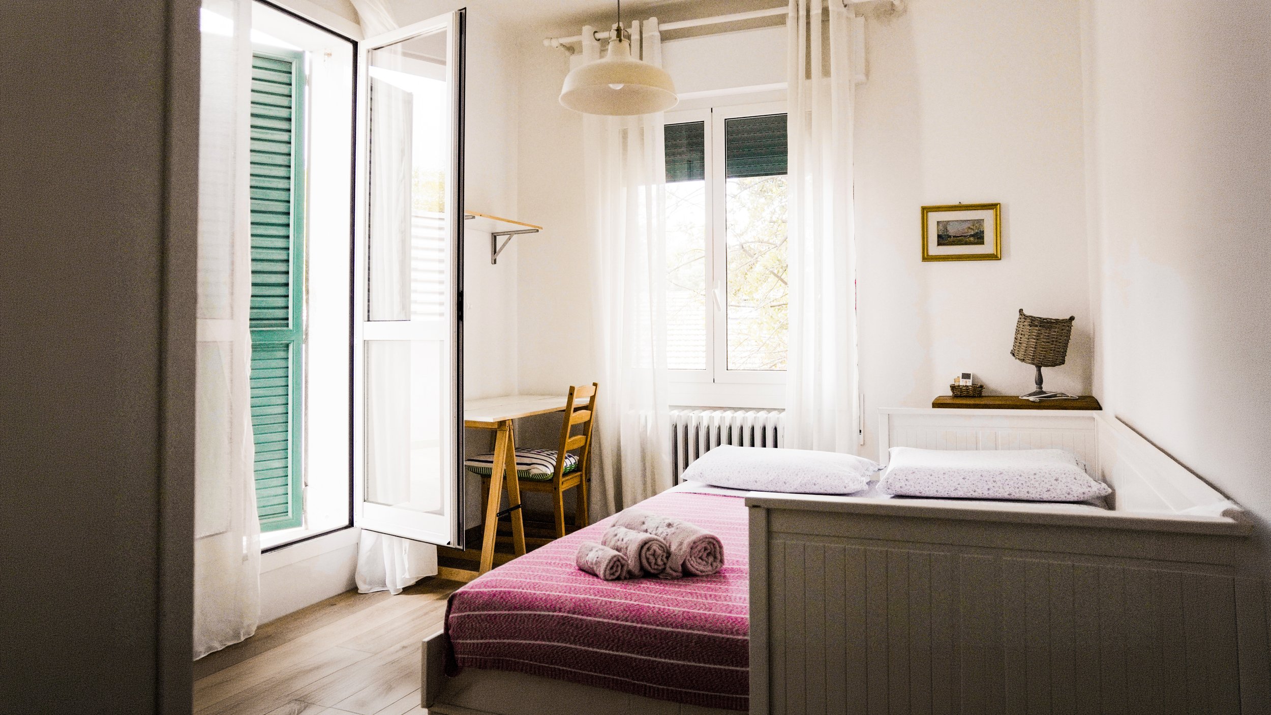 Stanza da letto con letto a due piazza, lenzuola e cuscini, finestra con tende bianche, scrivania con sedia, lampada da tavolo, decorazioni semplici.