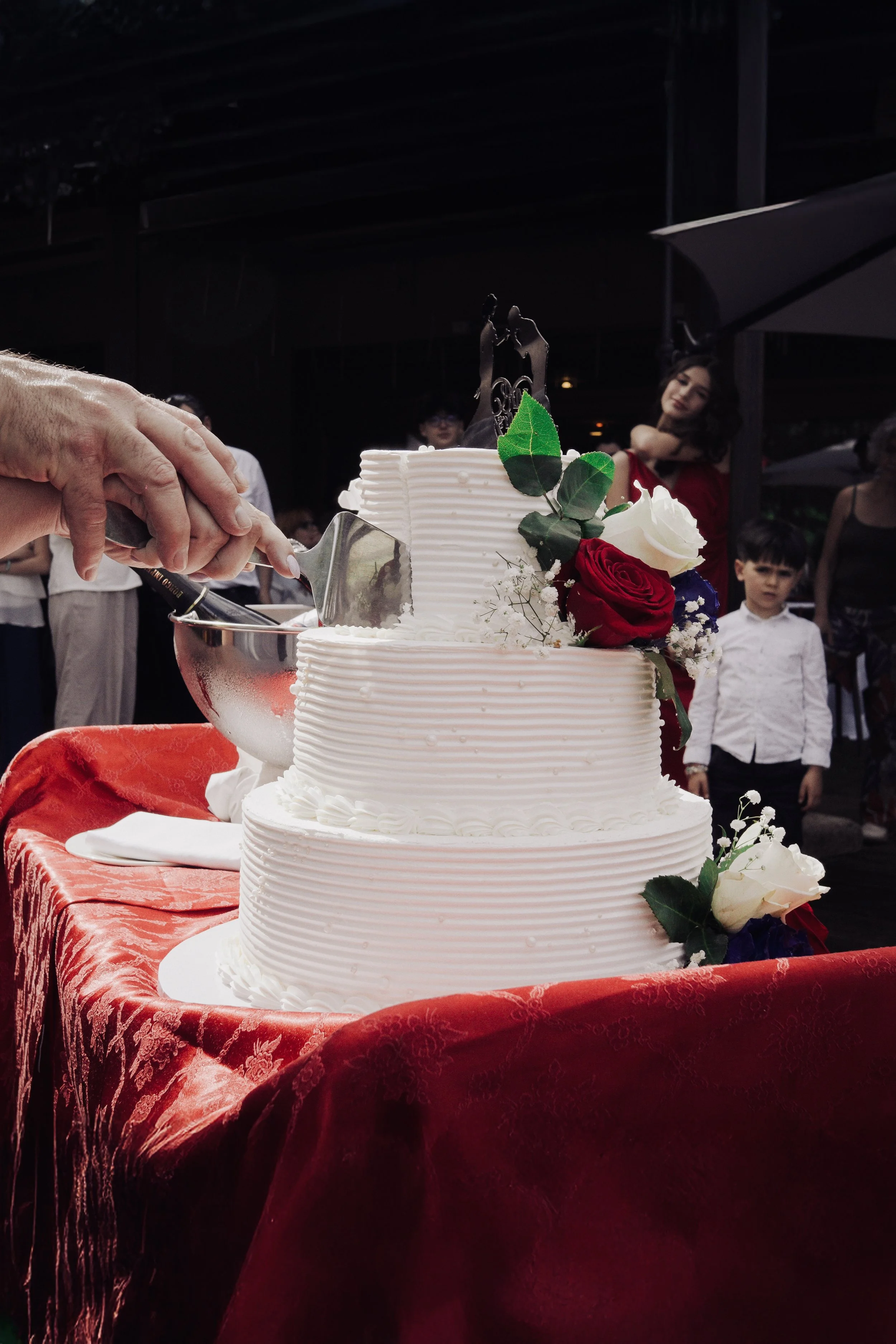 Torta di matrimonio bianca decorata con rose rosse, bianche e fiori di Baby's Breath, con qualcuno che la taglia, su tavolo ricoperto di tessuto rosso