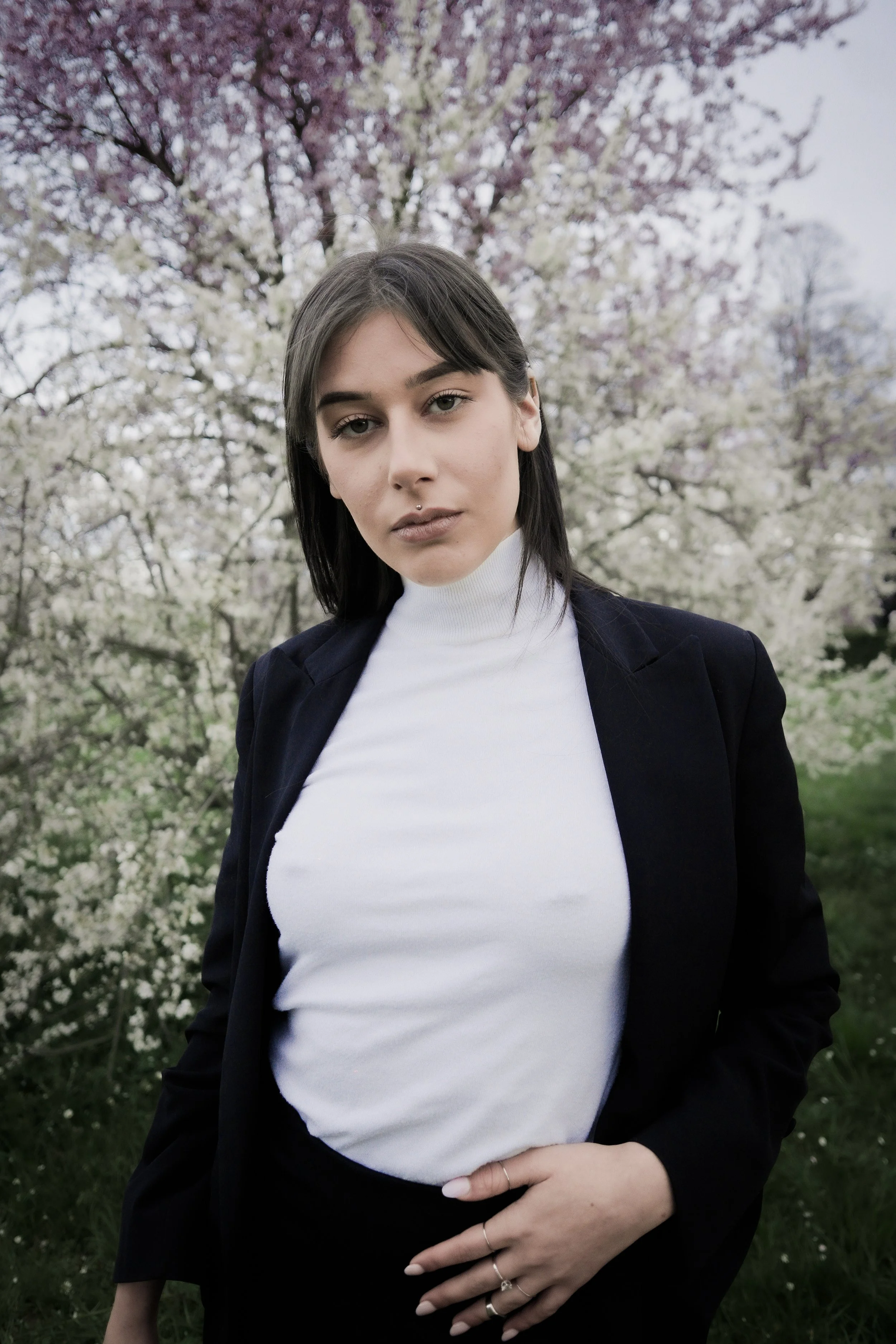 Una donna con capelli castani e occhi chiari, indossa una maglia bianca a collo alto e una giacca nera, si trova all'aperto con alberi in fiore rosa e bianco sullo sfondo.