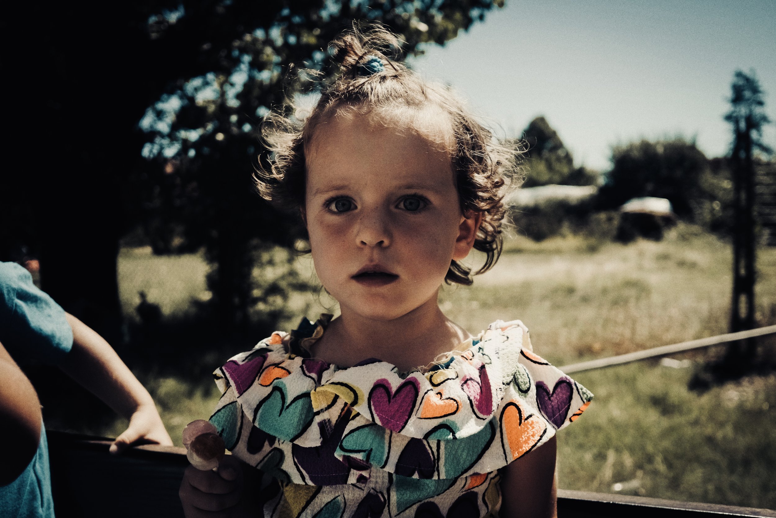 Ragazza con capelli riccioli e occhioni azzurri, vestita con un abito colorato a cuori, guarda verso la fotocamera in un ambiente all'aperto con alberi e cielo.