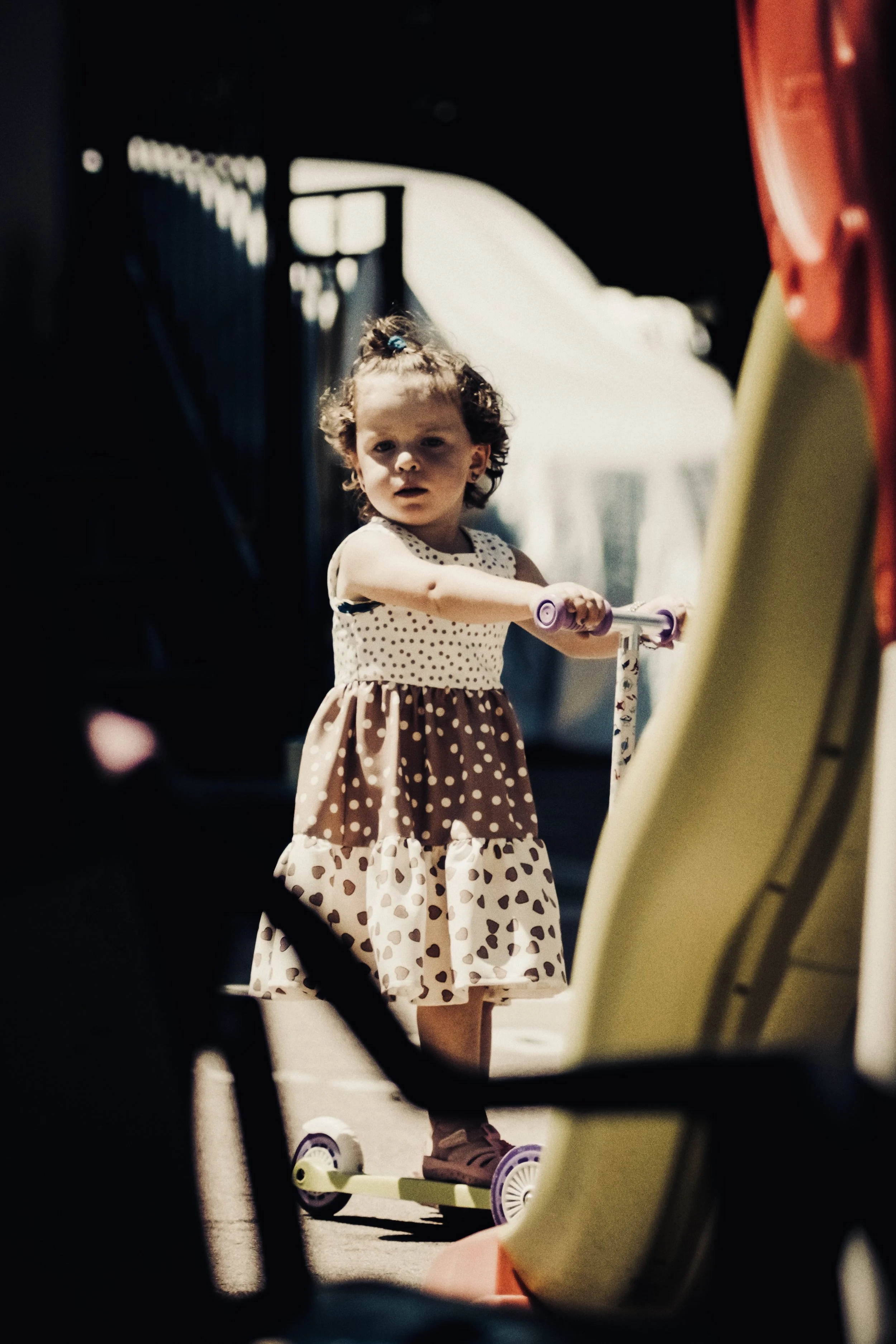 Una bambina con un vestito a pois ragazza piccola con scooter, che guarda verso la macchina fotografica, in un ambiente esterno.