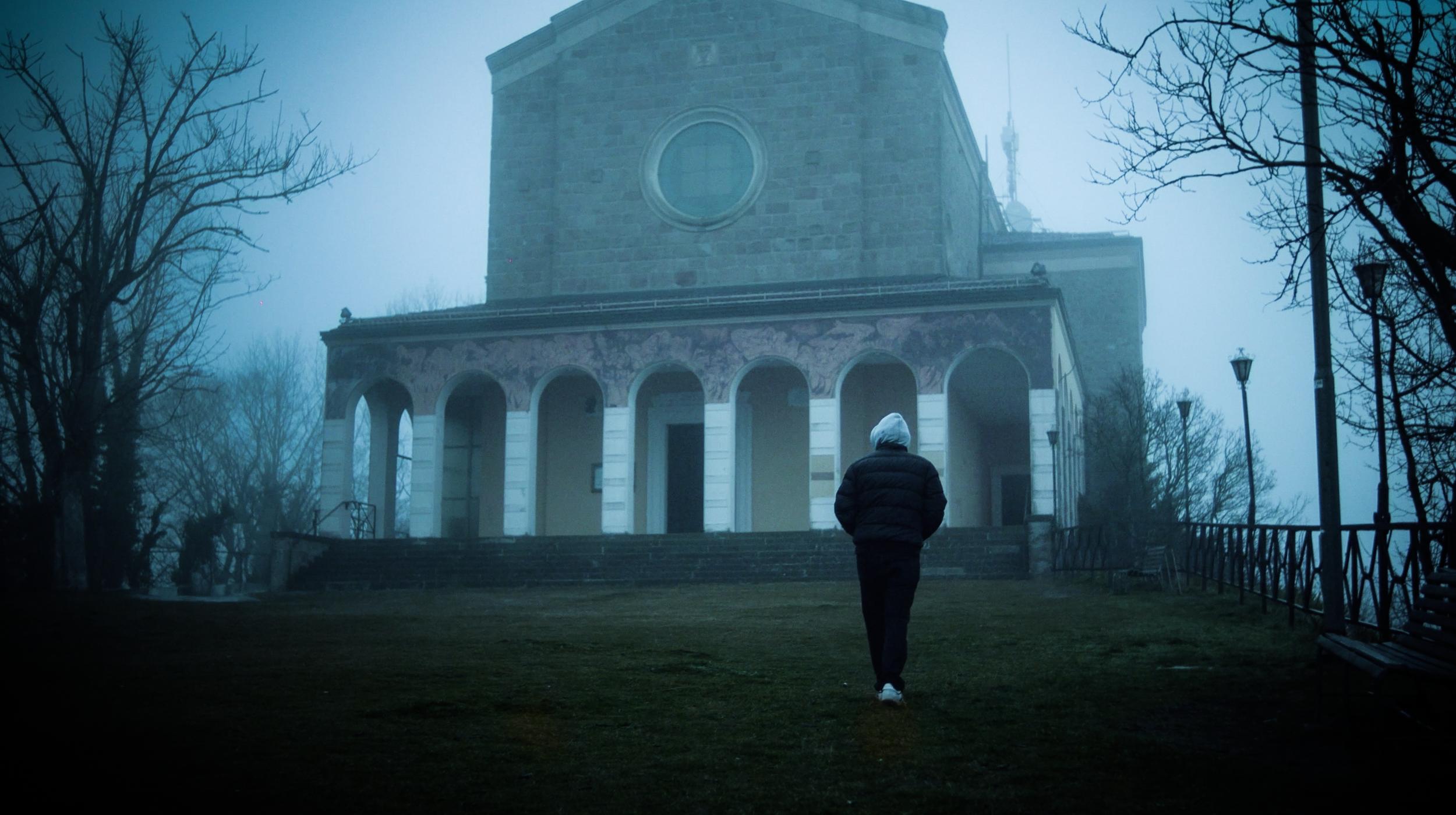 Una persona vestita con abiti pesanti cammina verso un edificio storico in condizioni di nebbia. L'edificio ha una struttura in pietra e un'entrata con archi. Il paesaggio circostante è innevato e ci sono alberi senza foglie ai lati.