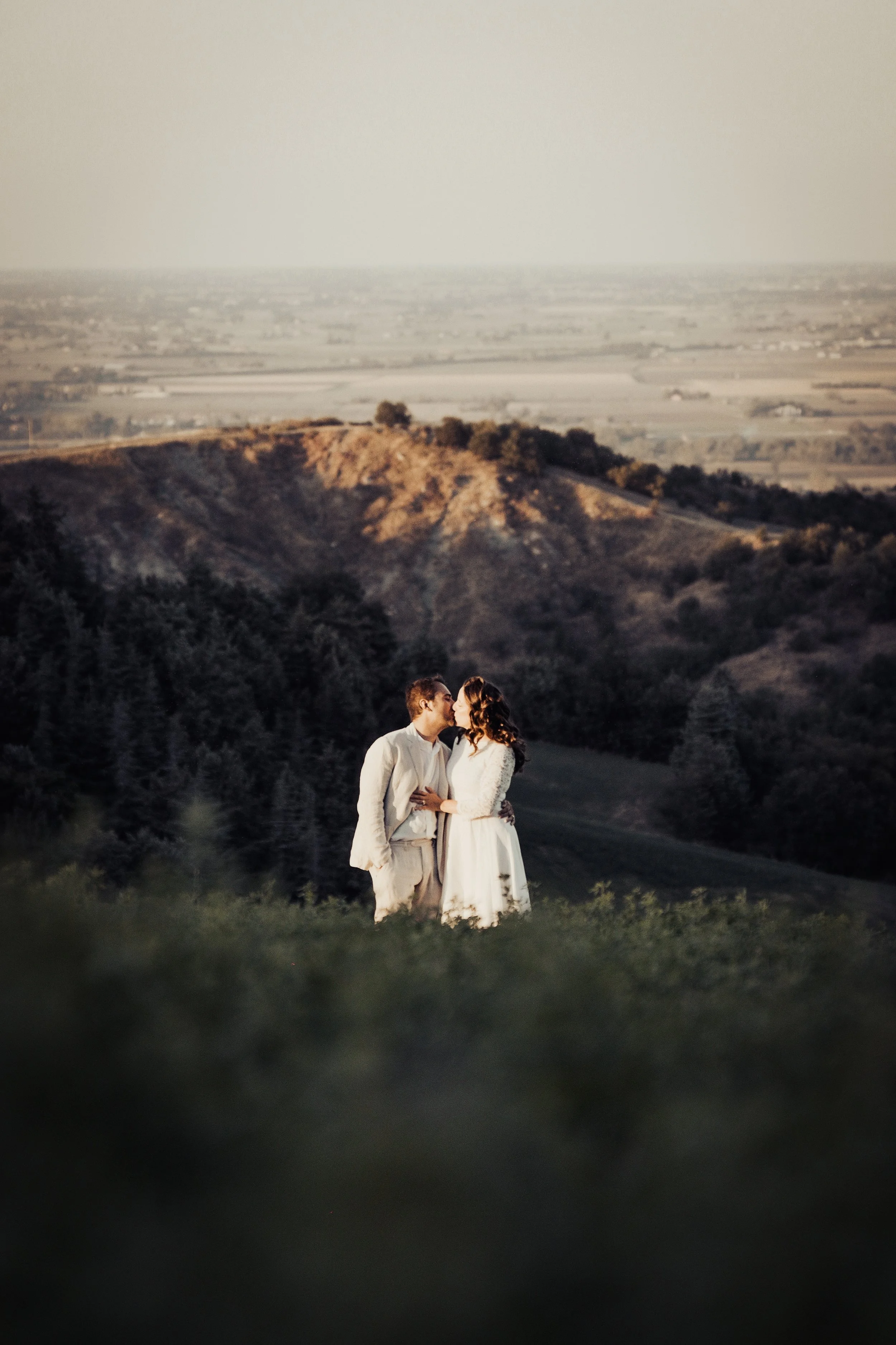Una coppia di sposi in abiti da matrimonio si bacia in un paesaggio collinare al tramonto, con campi e colline sullo sfondo.