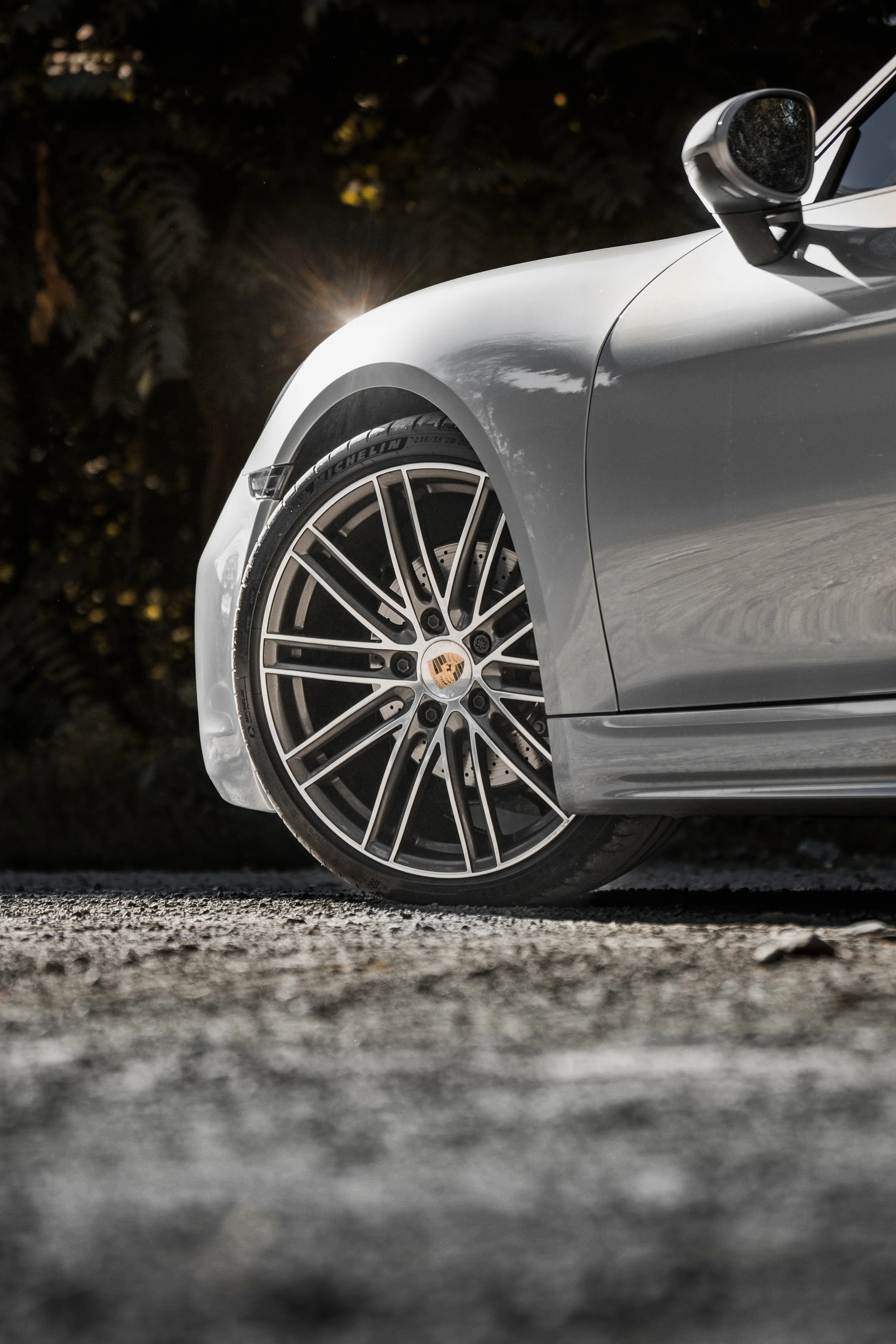 Metà anteriore di un'auto sportiva di colore argento, inquadratura laterale, con focus sulla ruota con rinforzo Porsche e pneumatico Michelin, su strada sterrata con sfondo di vegetazione.