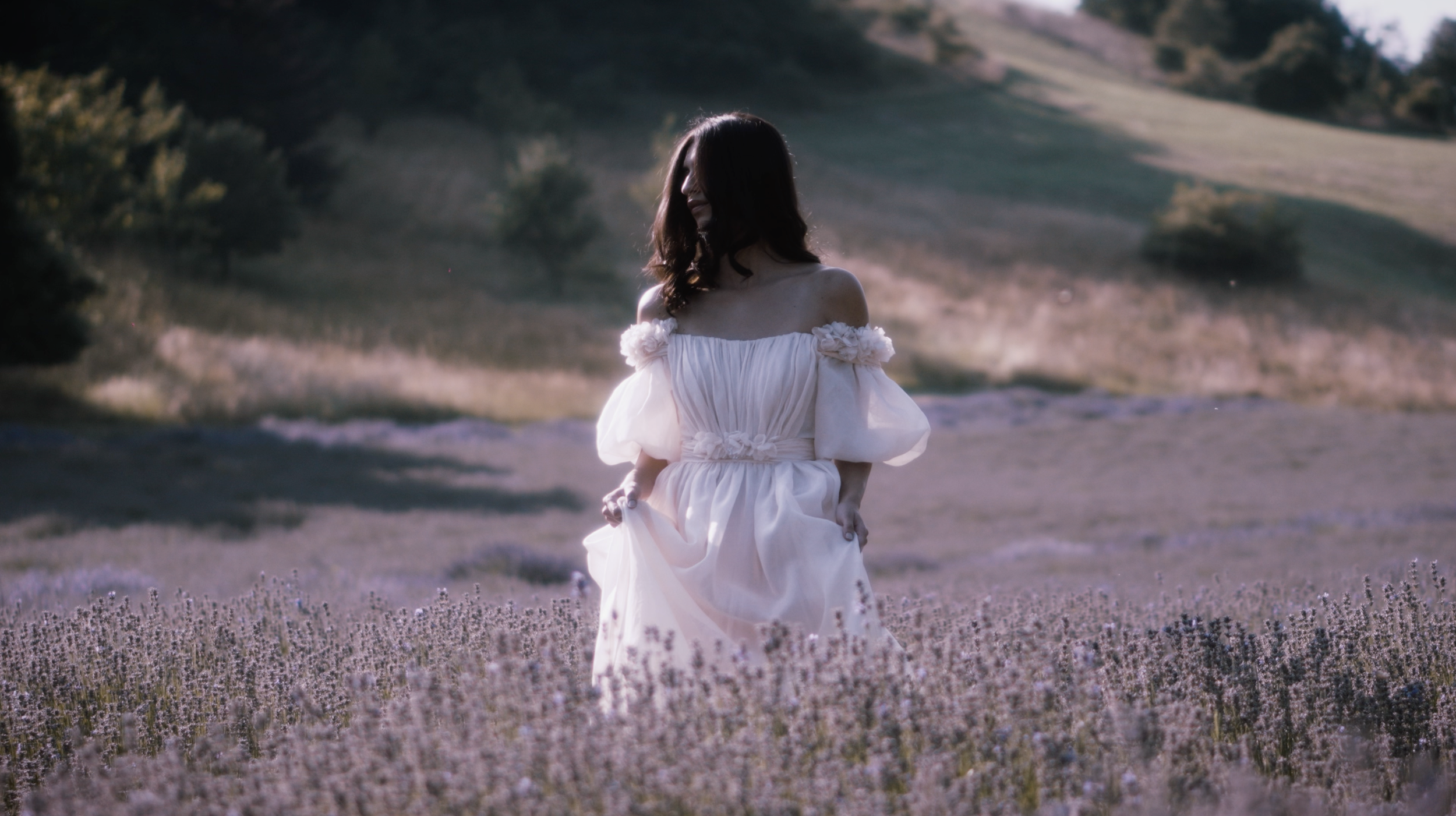 Donna in vestito bianco in un campo di lavanda con uno sfondo di colline verdi e alberi al tramonto.