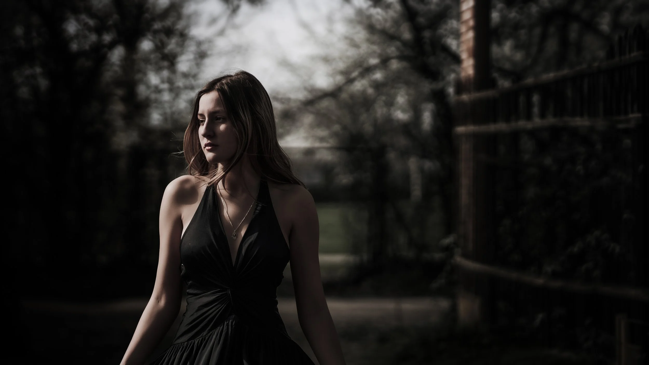 Ragazza con vestito nero e collana in un ambiente naturale con alberi e fitta ombra.