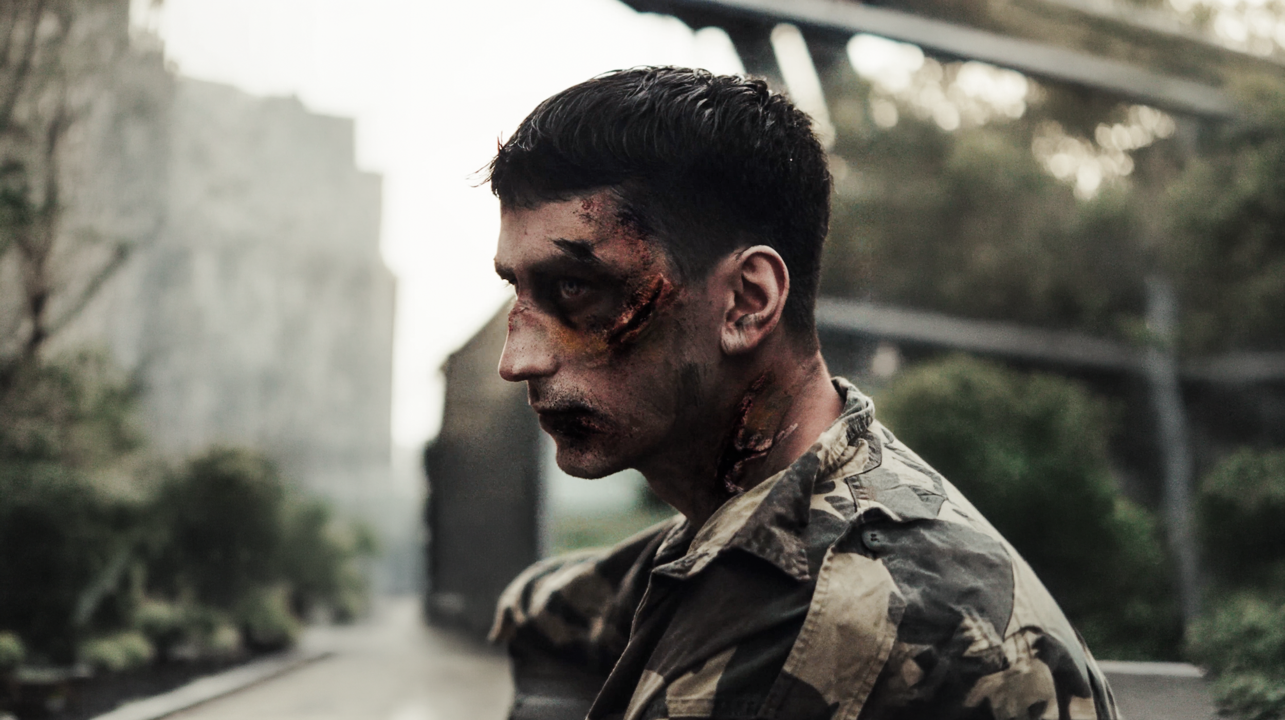 Uomo con ferite e graffi sul viso e collo, vestito con camicia mimetica, sangue e ferite visibili, ambiente urbano con structures in sfondo.