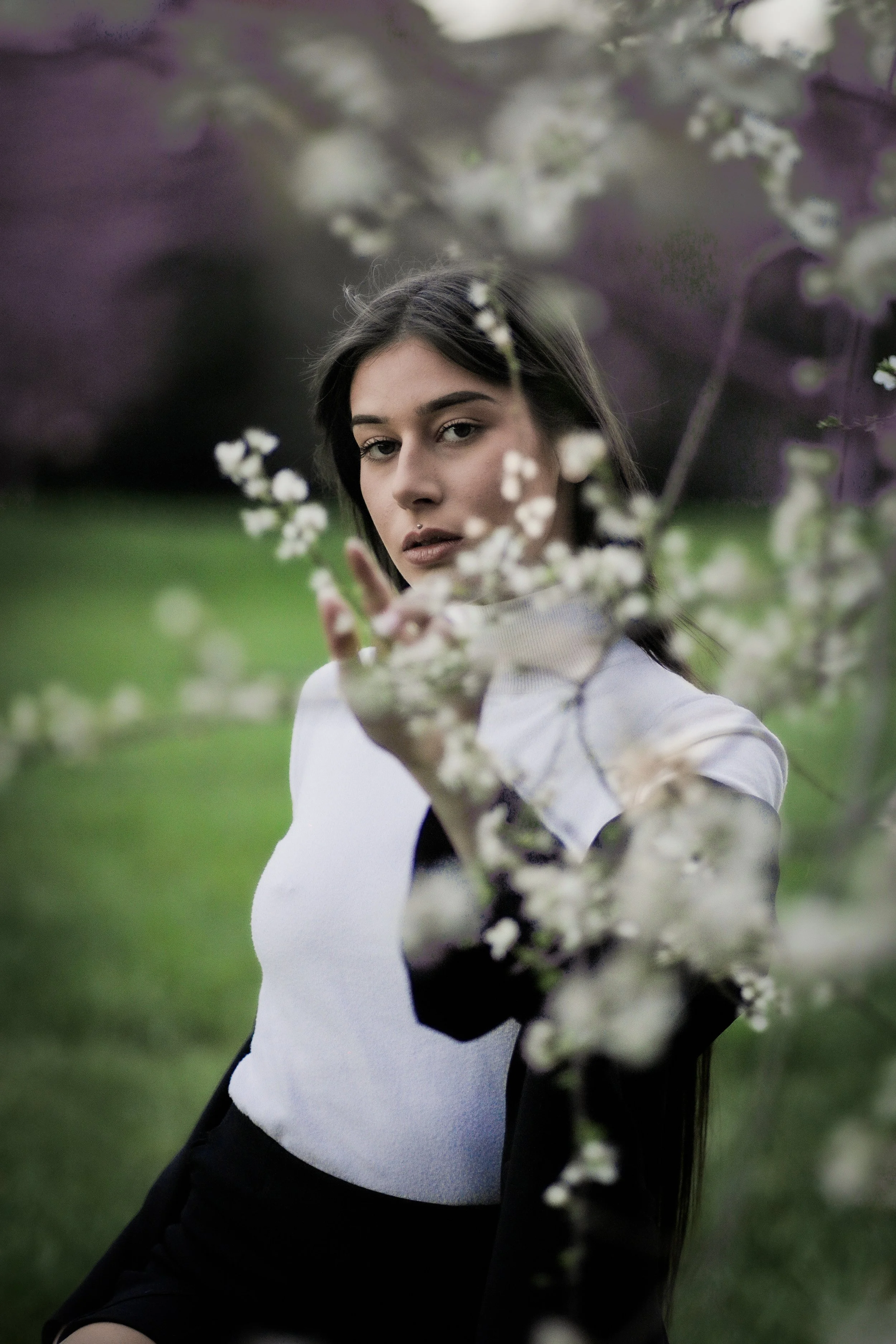 Una donna giovane tra i fiori di primavera si trova all'aperto, rivolta leggermente di lato, con un'espressione calma. Lo sfondo è verde e sfocato.