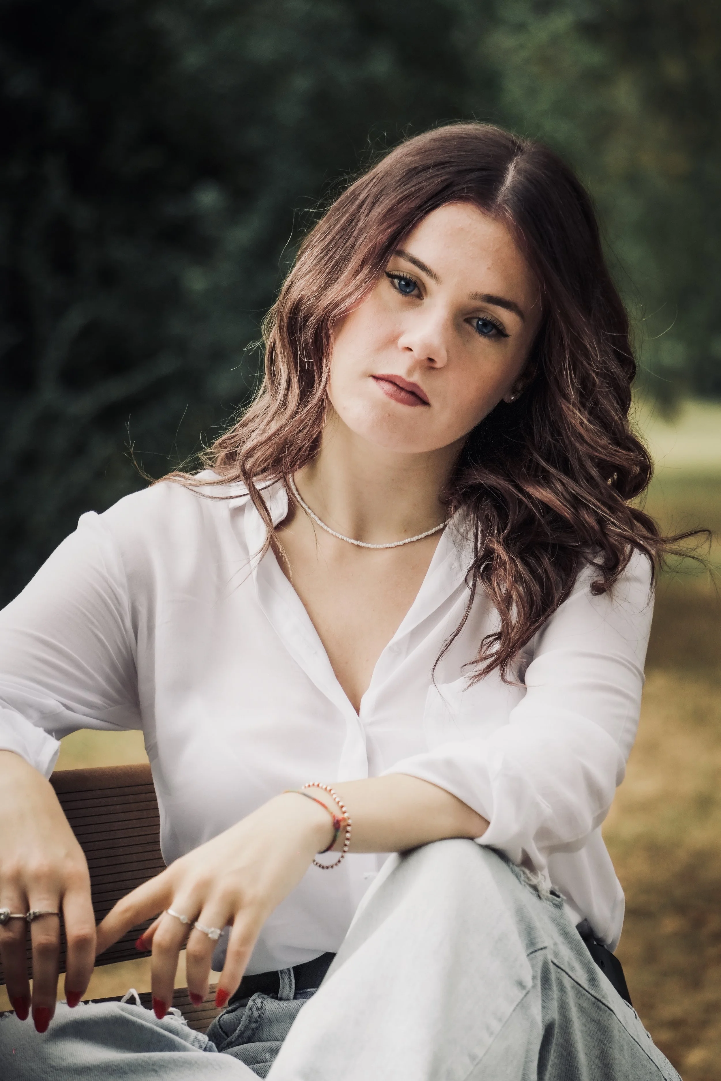 Ragazza con i capelli ondulati e cappelli chiari, vestita con camicia bianca, seduta su una panchina in un parco, con uno sfondo di alberi verdi.