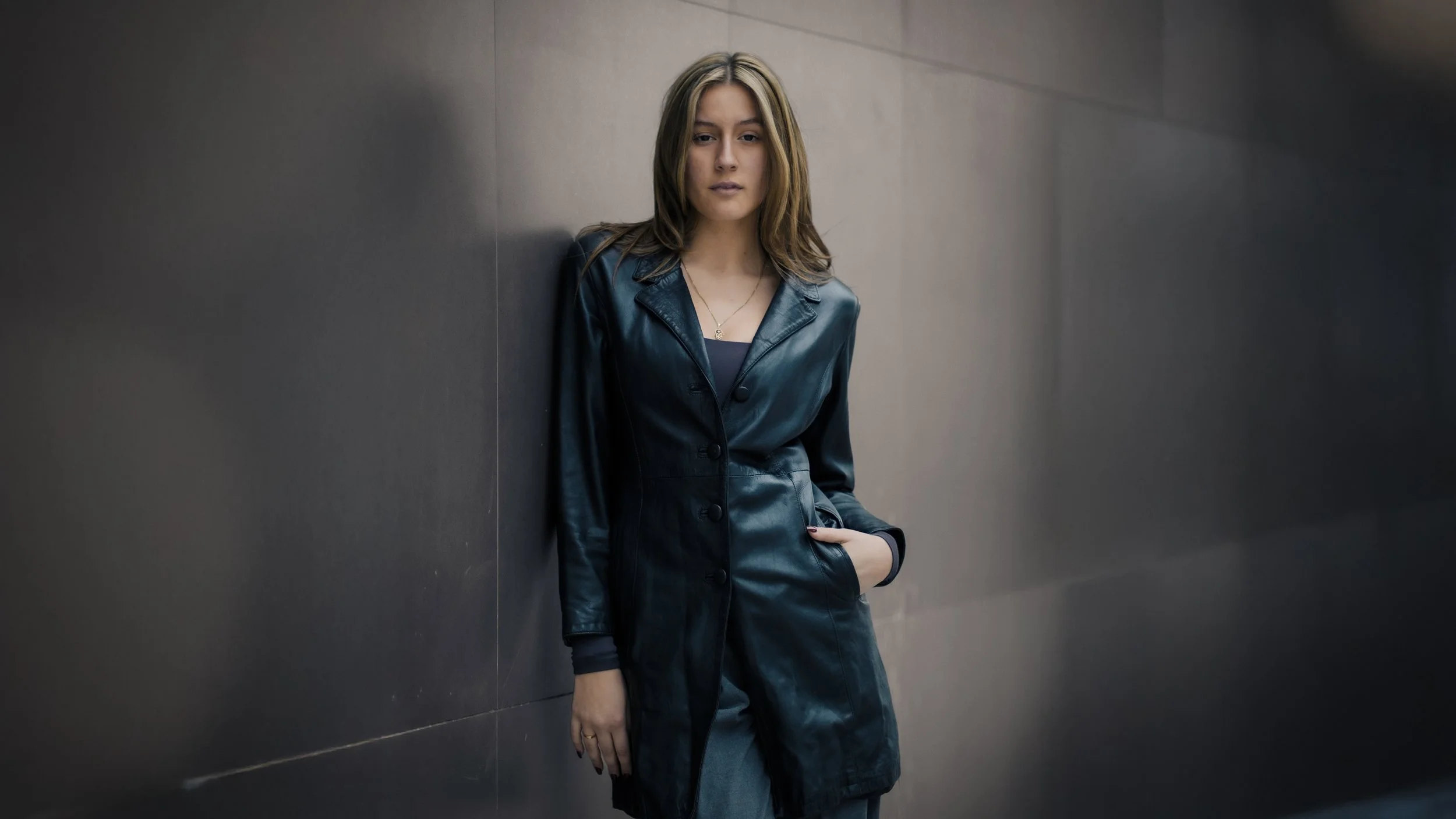 Una donna giovane con capelli castani e lunghi indossa un lungo cappotto di pelle blu scura e sta appoggiata a una parete di colore grigio scuro.