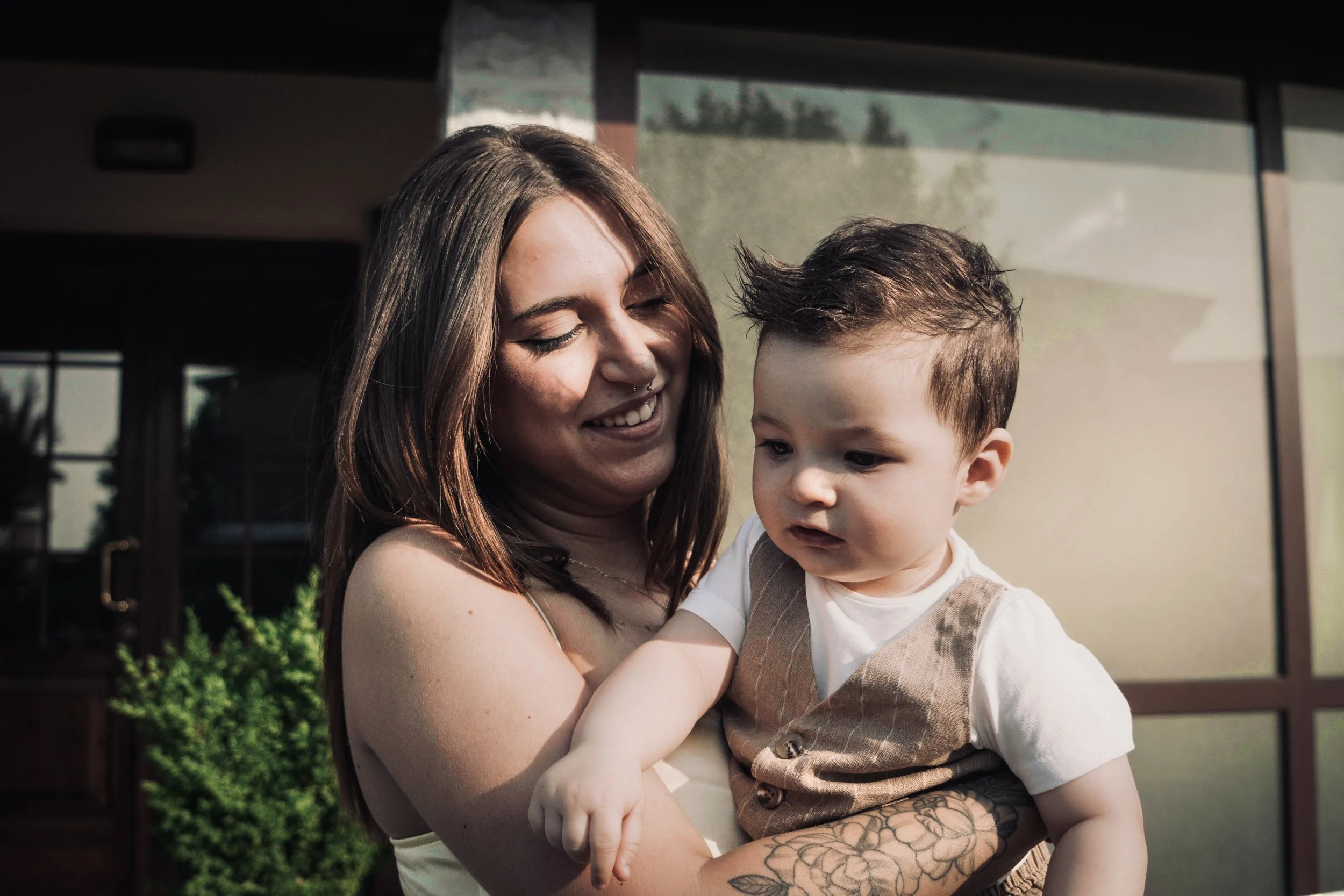 Una donna sorridente tiene in braccio un bambino, entrambi all'aperto davanti a una casa con grandi finestre.