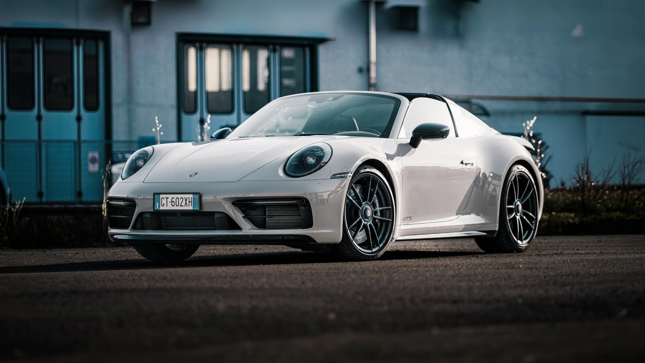 Auto Porsche Carrera cabrio bianca parcheggiato su strada, con edificio blu sullo sfondo.