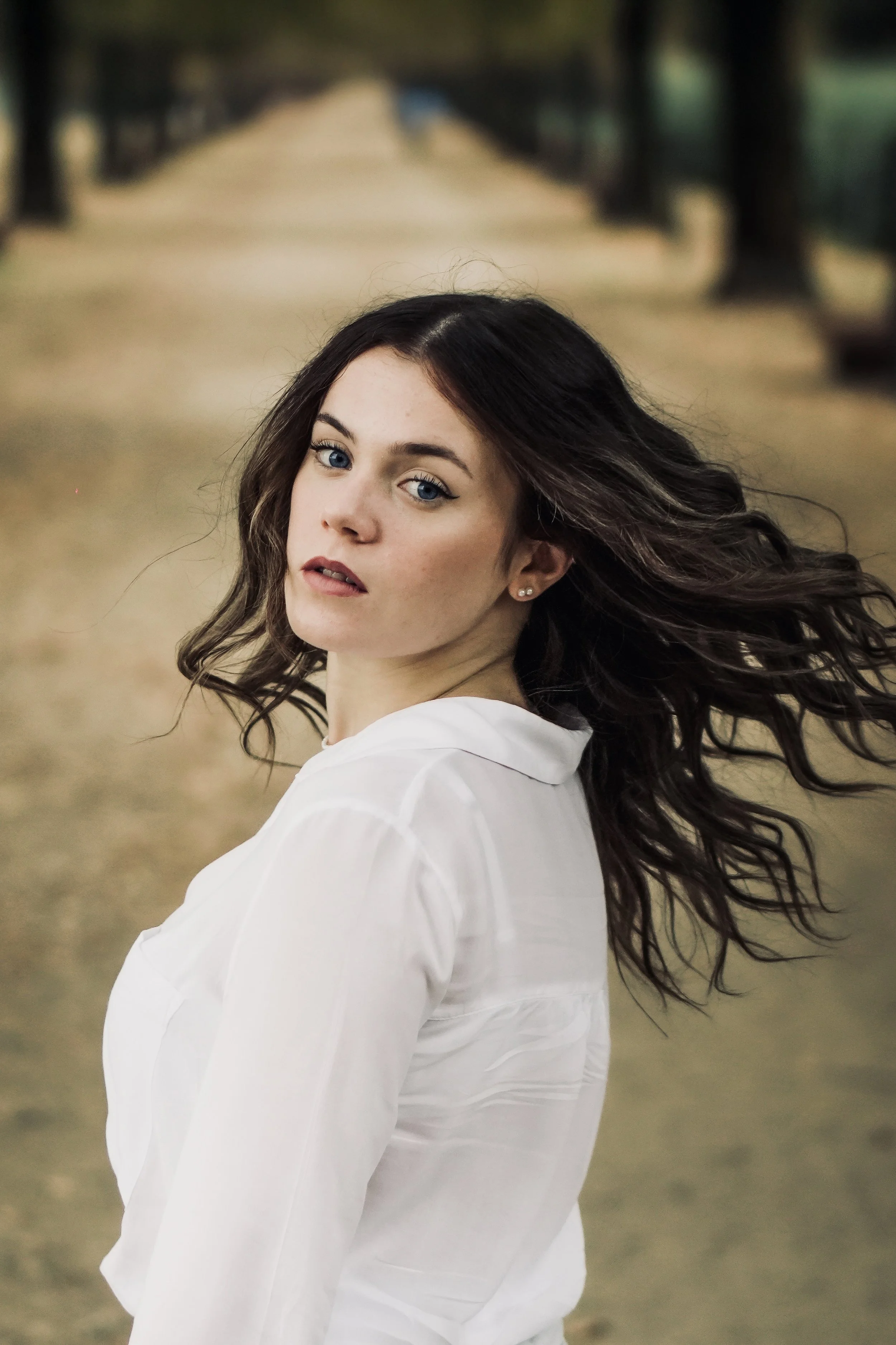 Ragazza con occhi azzurri e capelli castani mossi, che indossa una camicia bianca, che guarda verso la fotocamera, all'aperto su una strada alberata.