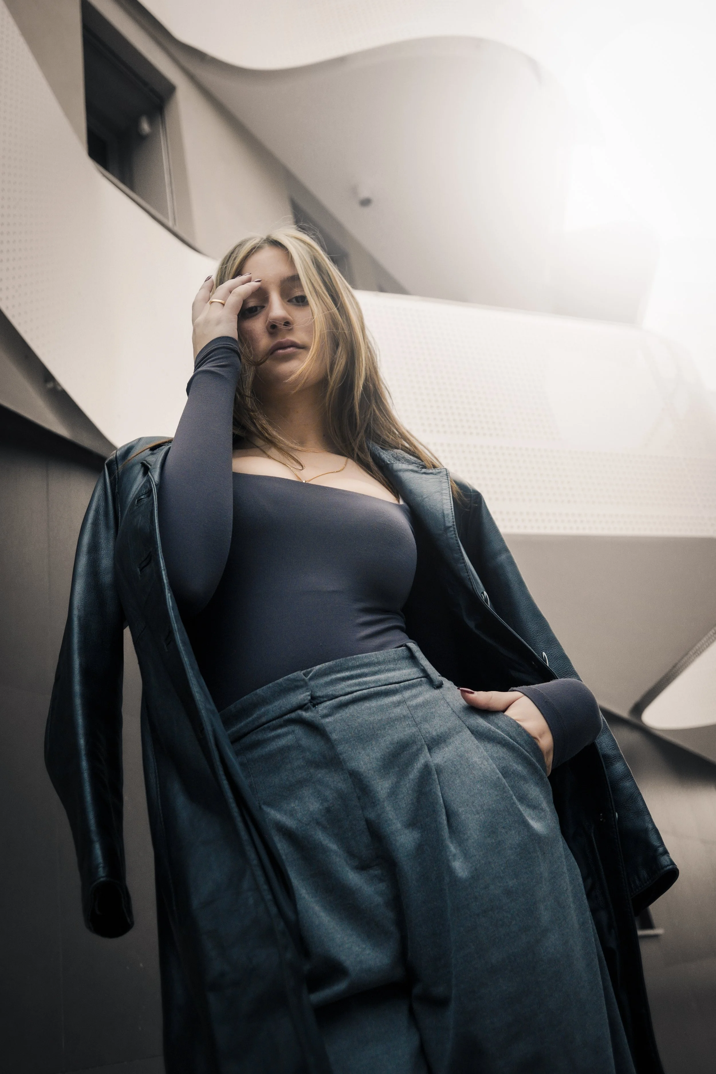 Una donna giovane indossa abiti scuri e si appoggia a un muro moderno, con un'espressione pensierosa.