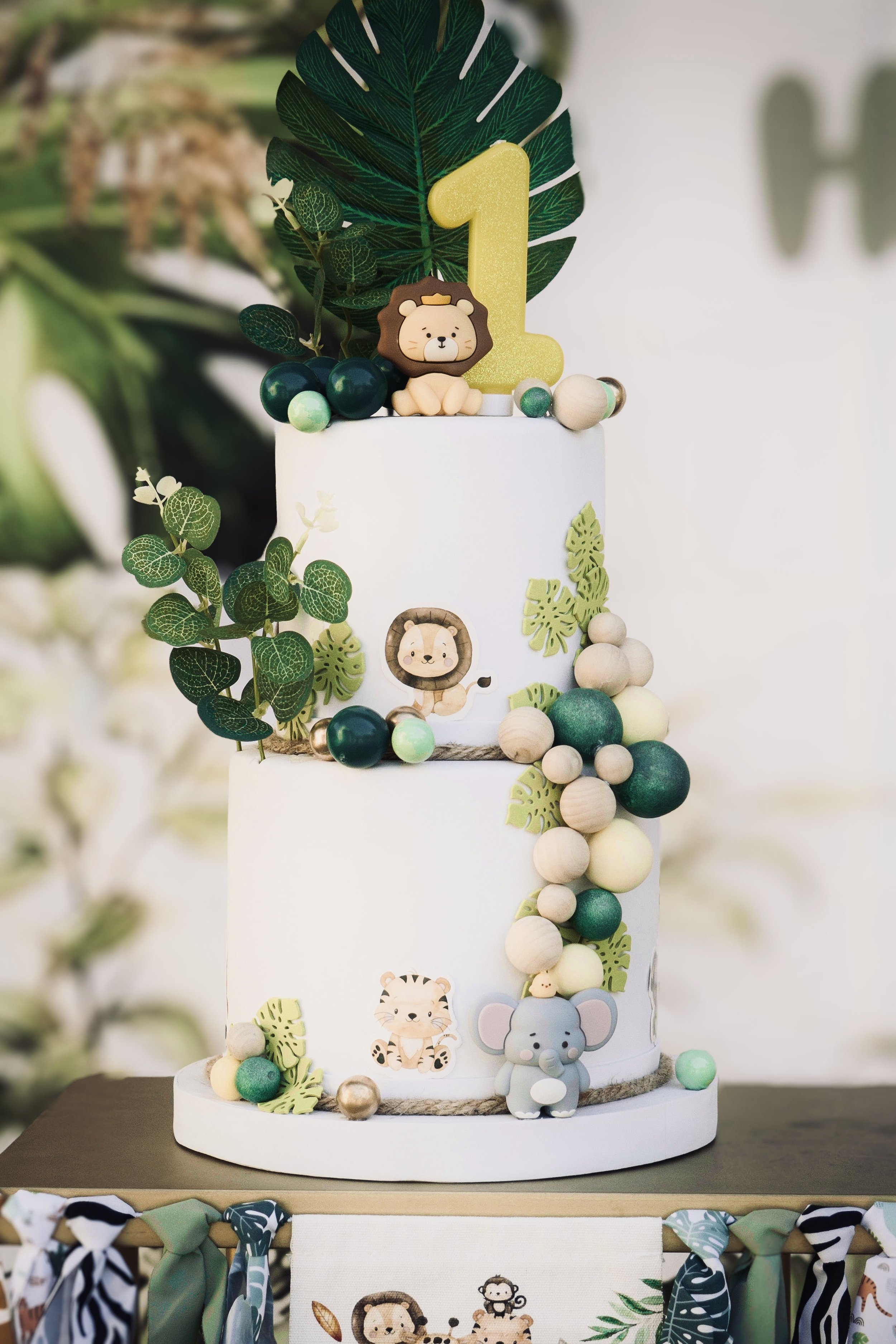 Torta di compleanno a tre piani decorata con tema della giungla, con animali di carta e fiori, e un grande numero 1 dorato in cima, circondata da foglie verdi e palline decorative