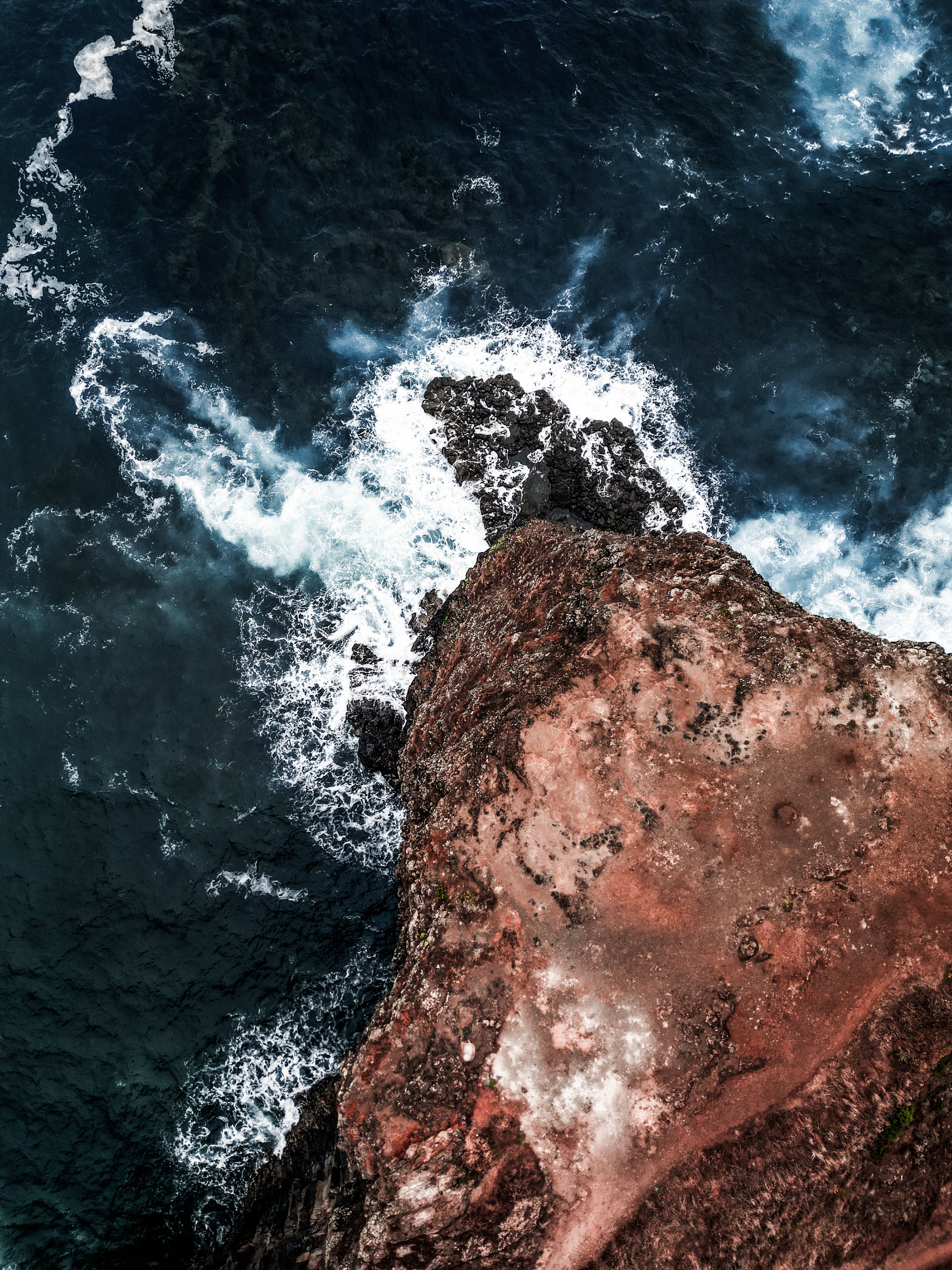 Vista dall'alto di una scogliera rossa che si affaccia sul mare, con onde che si infrangono contro le rocce.