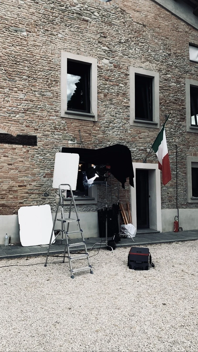 Setup di riprese fotografiche o video all'esterno di un edificio con parete in mattoni, bandiera italiana, luci e attrezzature di scena.