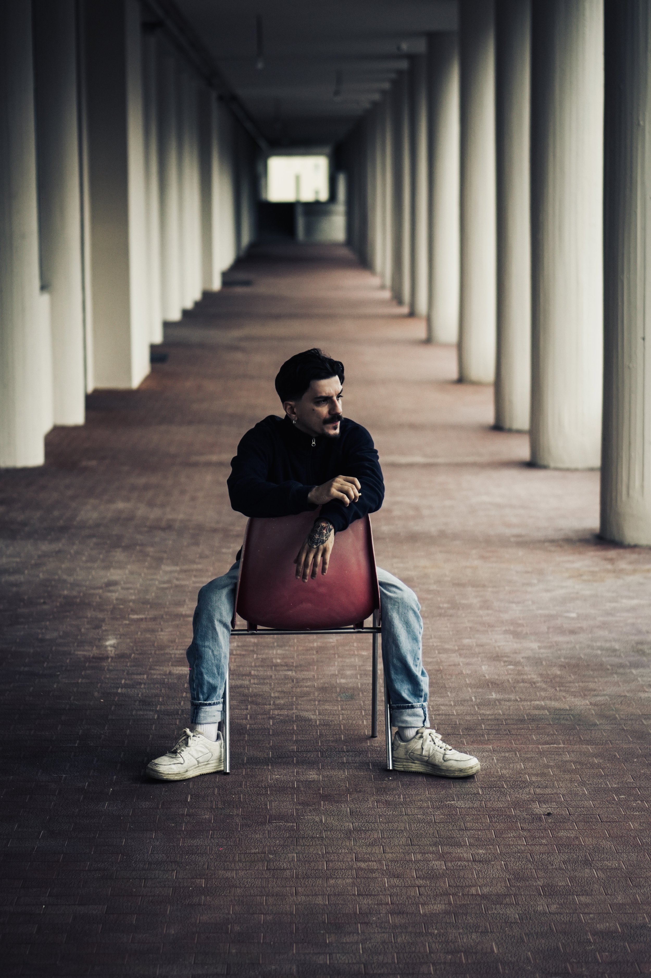 Persona seduta su una sedia rossa in un corridoio lungo con colonne, con espressione pensierosa.