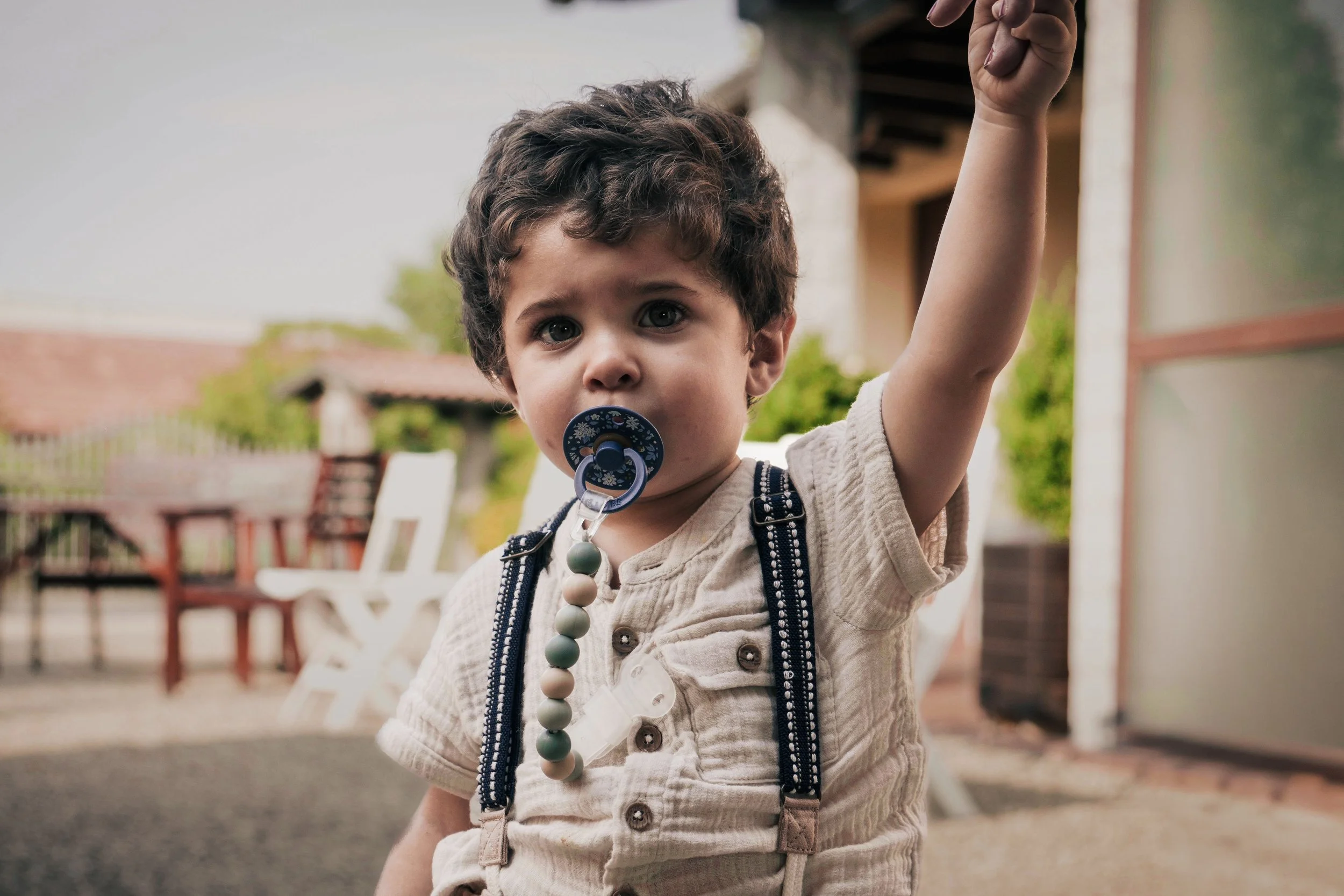 Un bambino con i capelli castani ricci, indossa una maglietta beige e una tutina, con bretelle blu con pois bianchi, tiene un ciuccio con una catena di perle color pastello, e alza il braccio destro mentre guarda verso la fotocamera, sullo sfondo un ambiente esterno con tavoli e sedie di giardino.