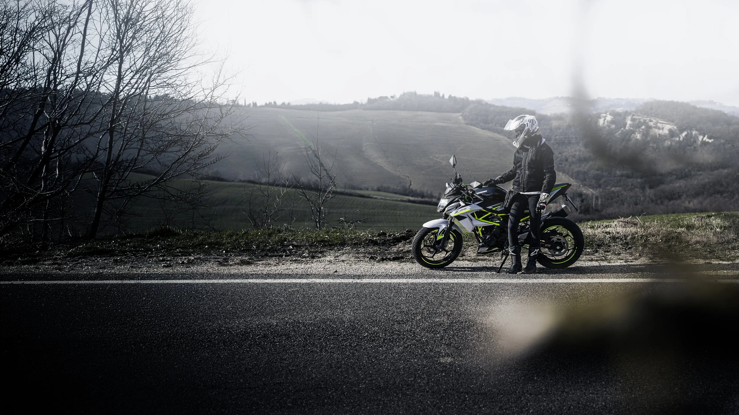 Un motociclista in abbigliamento protettivo vicino a una moto da strada su una strada di campagna con paesaggi collinari e cielo nuvoloso.