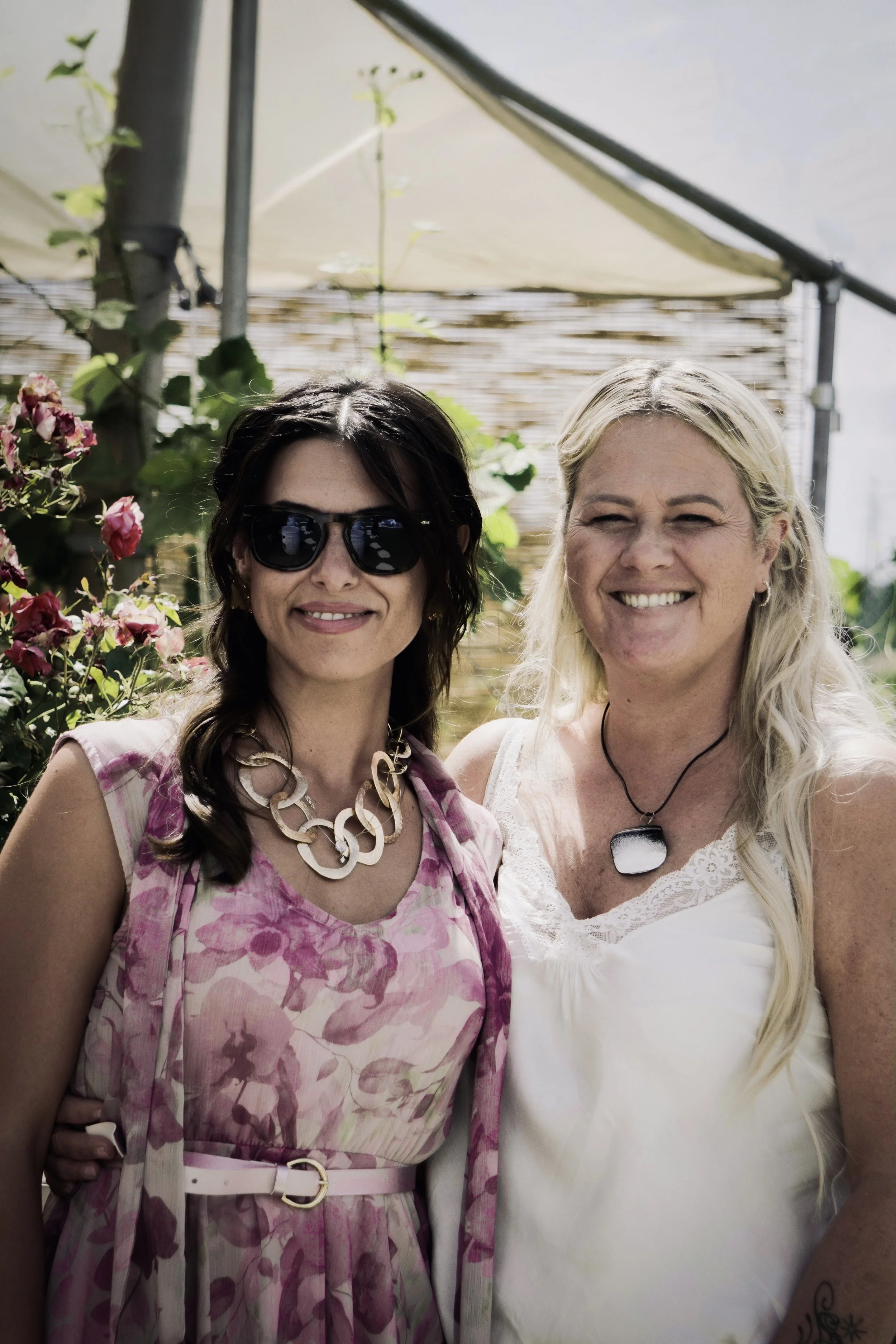 Due donne sorridenti, una con occhiali da sole e l'altra con capelli biondi, si trovano in un ambiente all'aperto circondate da piante e fiori.