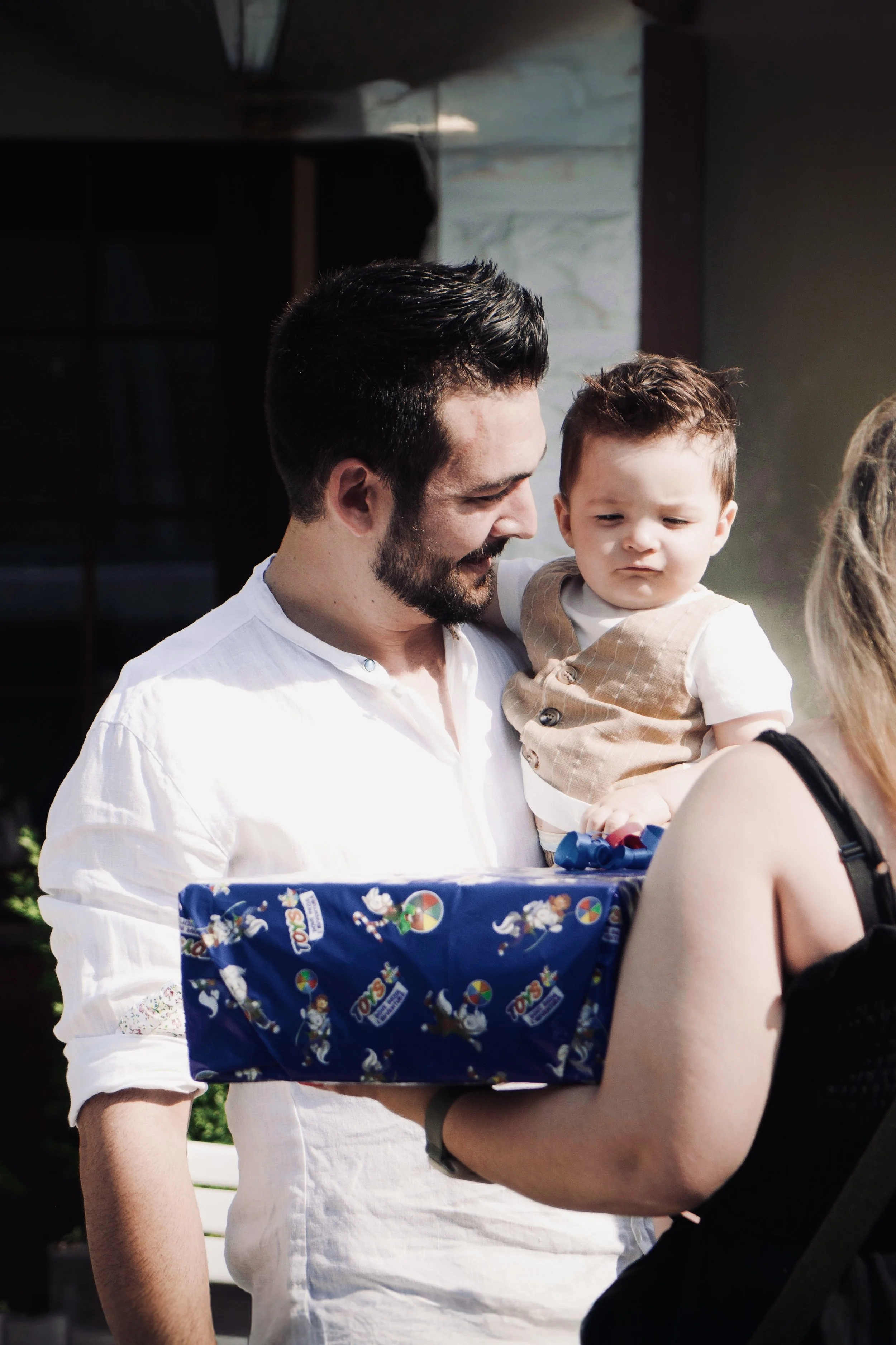 Un uomo che sorride mentre tiene un bambino in braccio. La donna in primo piano tiene un regalo avvolto in carta blu con disegni di cavalli e carretti.