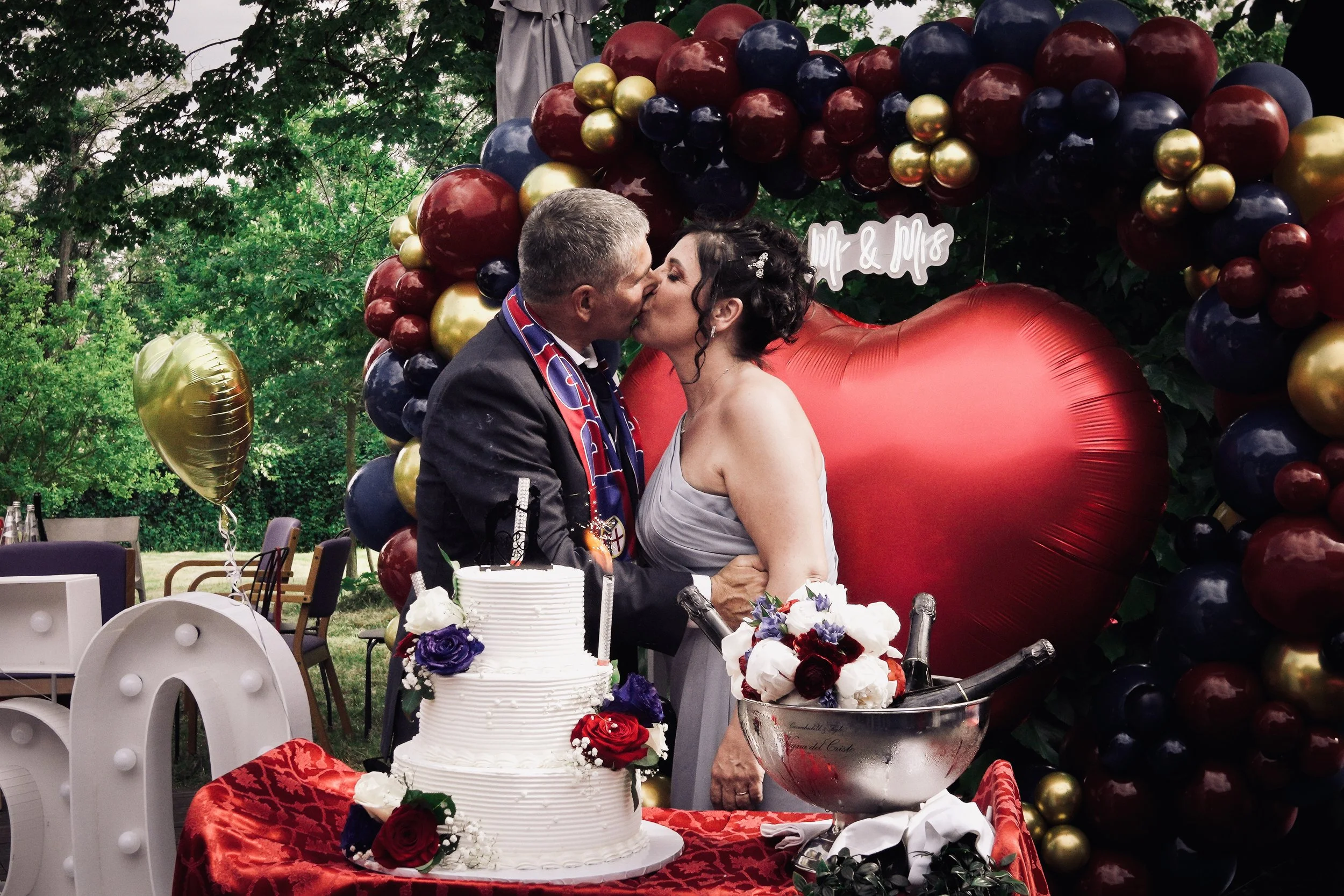 Una coppia di sposi si bacia durante il matrimonio, circondati da decorazioni con palloncini rossi, blu e oro e un grande cuore rosso, con una torta bianca decorata con fiori rossi e viola.
