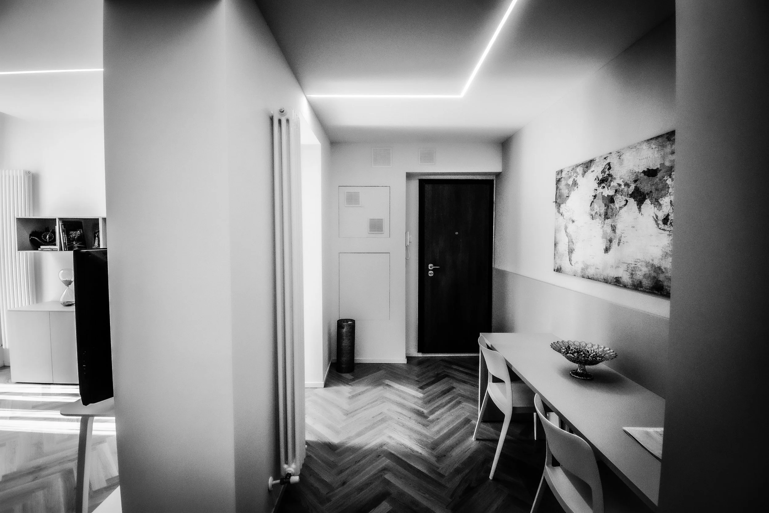 Interno di una casa moderna con pareti bianche, soffitto con luci a LED, pavimento in legno a spina di pesce, una porta nera, un tavolo bianco con sedie e un dipinto sulla parete.