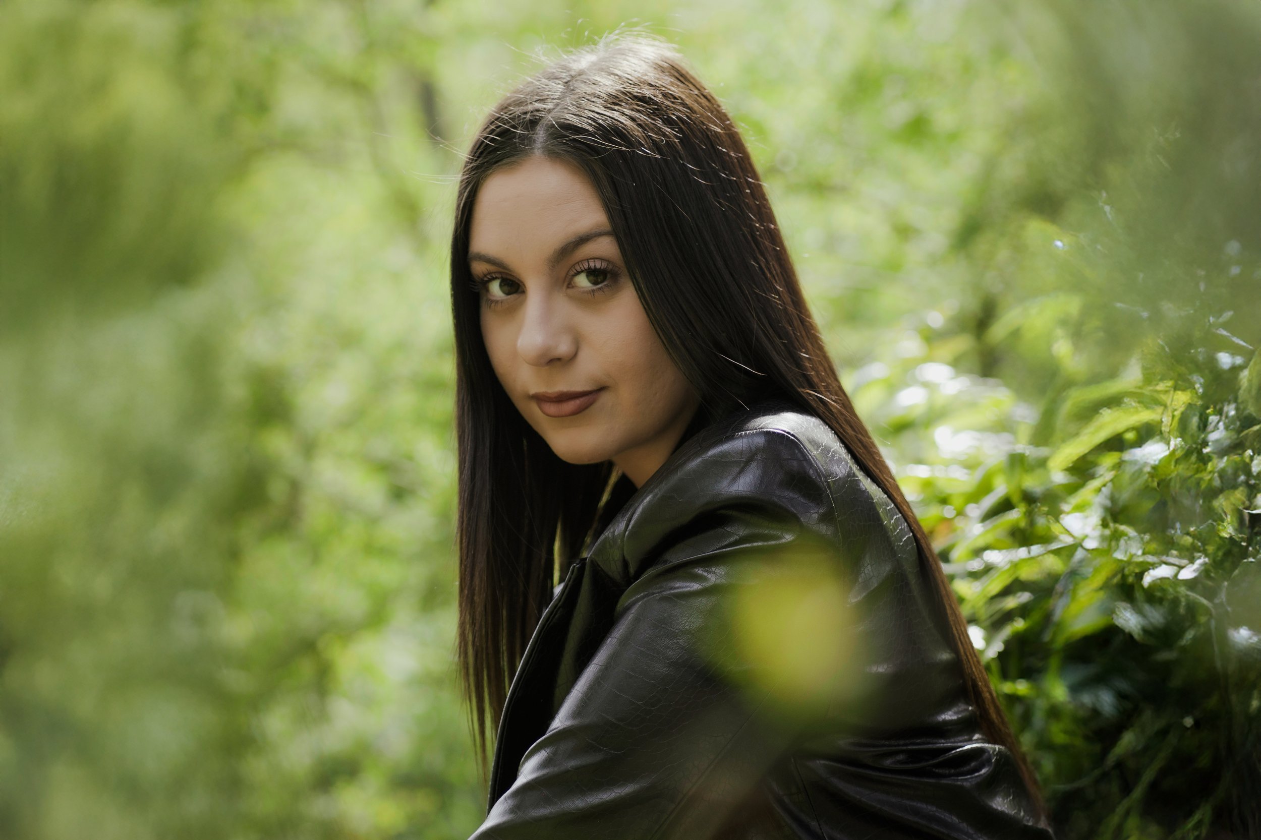 Una giovane donna con capelli lunghi e scuri, indossa una giacca di pelle nera, si trova all'aperto tra la vegetazione verde, guarda verso la fotocamera con un sorriso gentile.