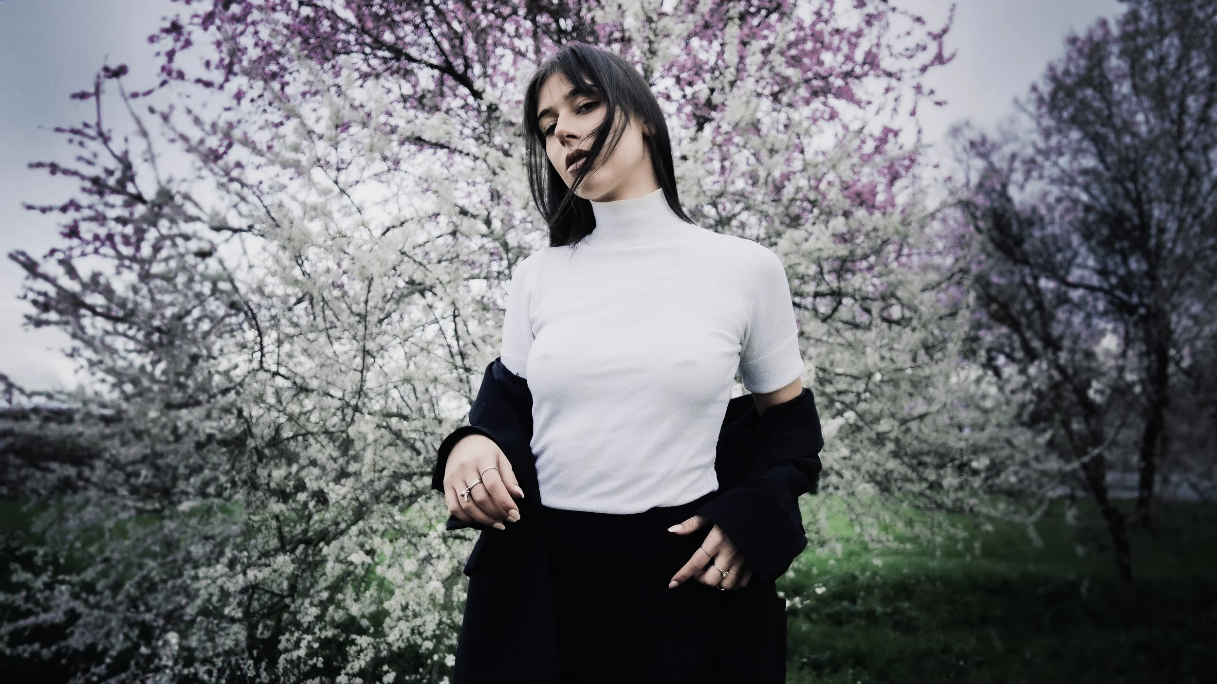 Una donna con capelli neri e vestiti scuri davanti a un albero di fiori di ciliegio in primavera