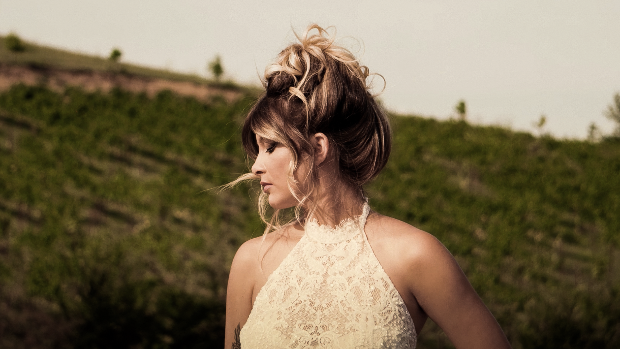 Donna con capelli riccioli e updo, indossa un vestito di pizzo beige, sfondo di colline verdi e arbusti.