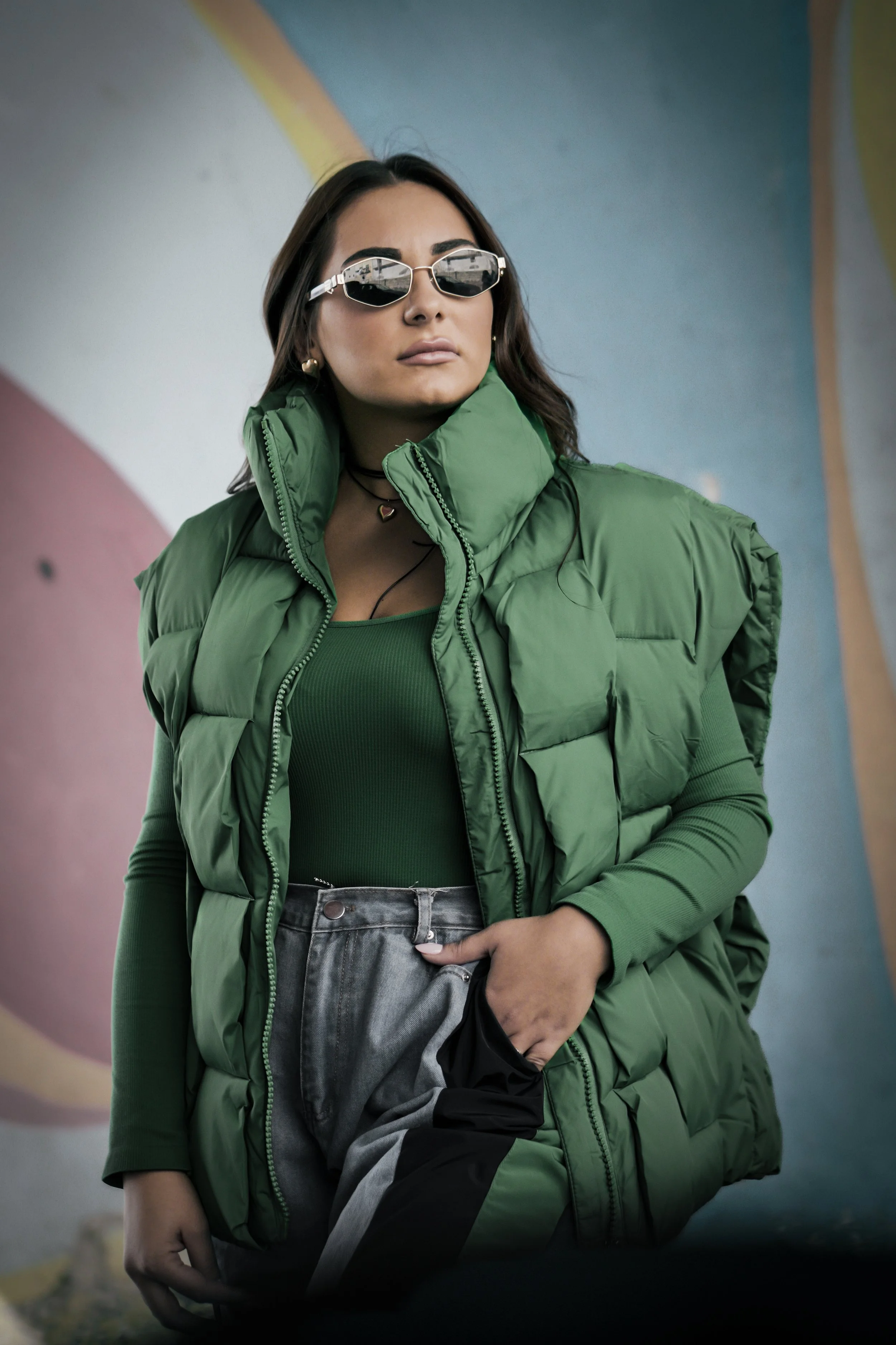Una donna con capelli castani lunghi, occhiali da sole e un abbigliamento verde, che indossa una giacca trapuntata verde, una maglietta verde a maniche lunghe, e pantaloni grigi, si trova davanti a un muro colorato con un arcobaleno dipinto.