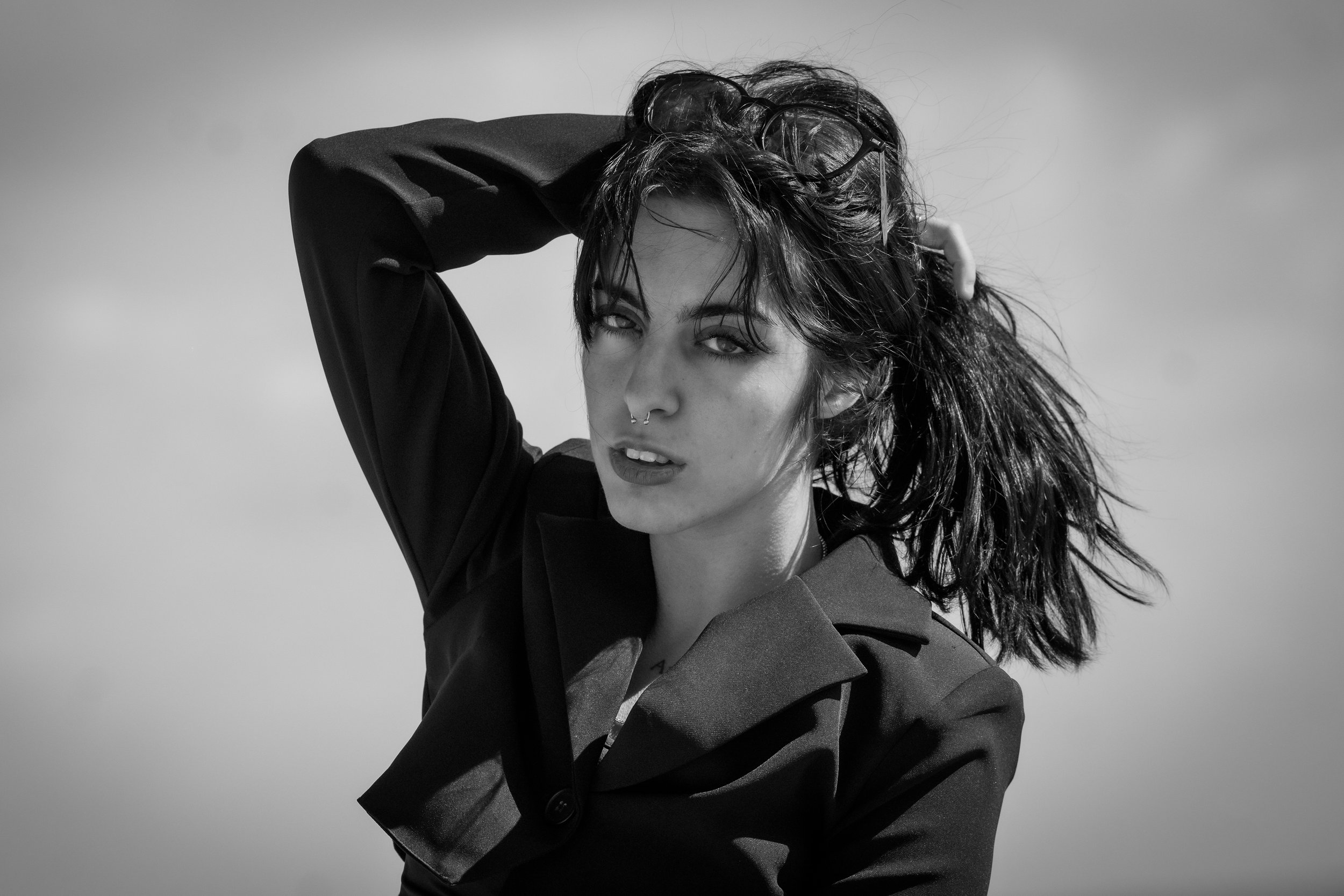 Ragazza con capelli spettinati, occhiali sulla testa, espressione seducente, indossa una giacca nera, sfondo chiaro, fotografia in bianco e nero.