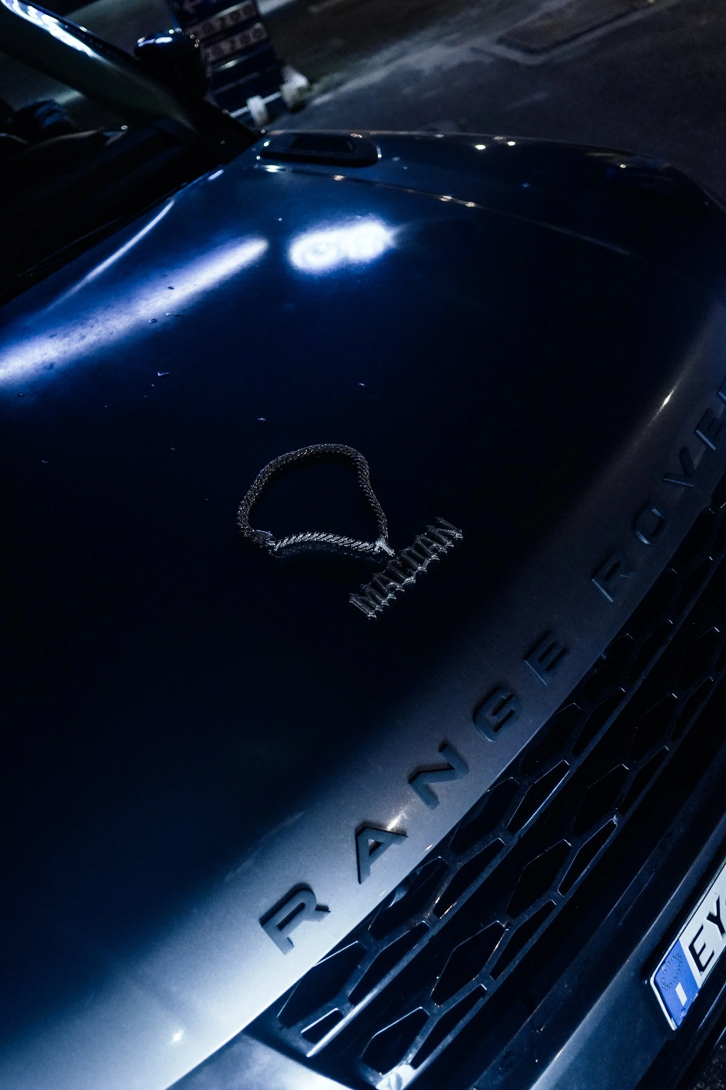 Il cofano di un'auto Rover Defender con un ciondolo e una collana sopra, serigrafata con la scritta 'RANGE ROVER', e un'auto a destra con targa europea.