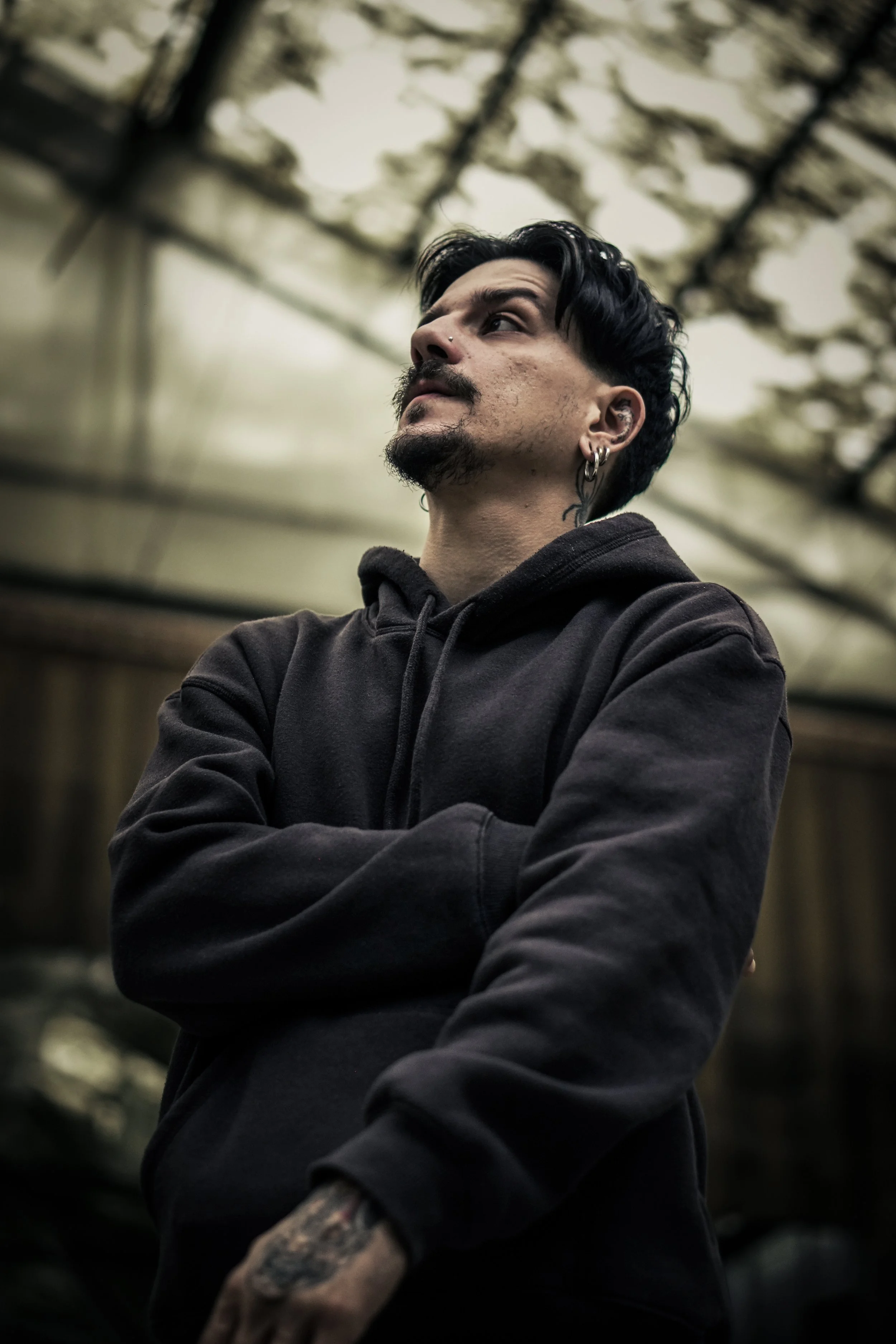 ritratto di un uomo con i capelli neri e barba, indossa felpa nera, con orecchini, stanco e pensieroso, in un ambiente chiuso con soffitto in vetro