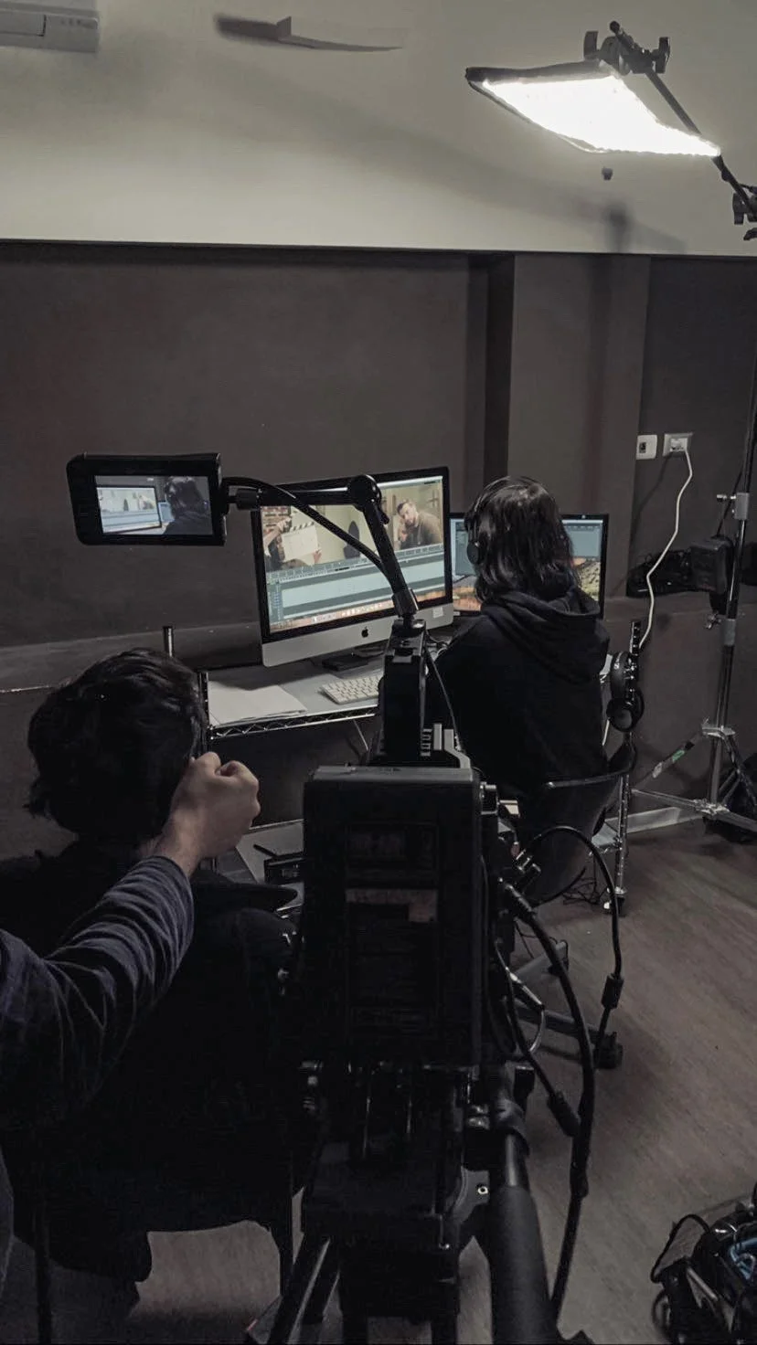 Studio di produzione video con due persone davanti a computer con software di editing, telecamere, luci e attrezzature di registrazione.